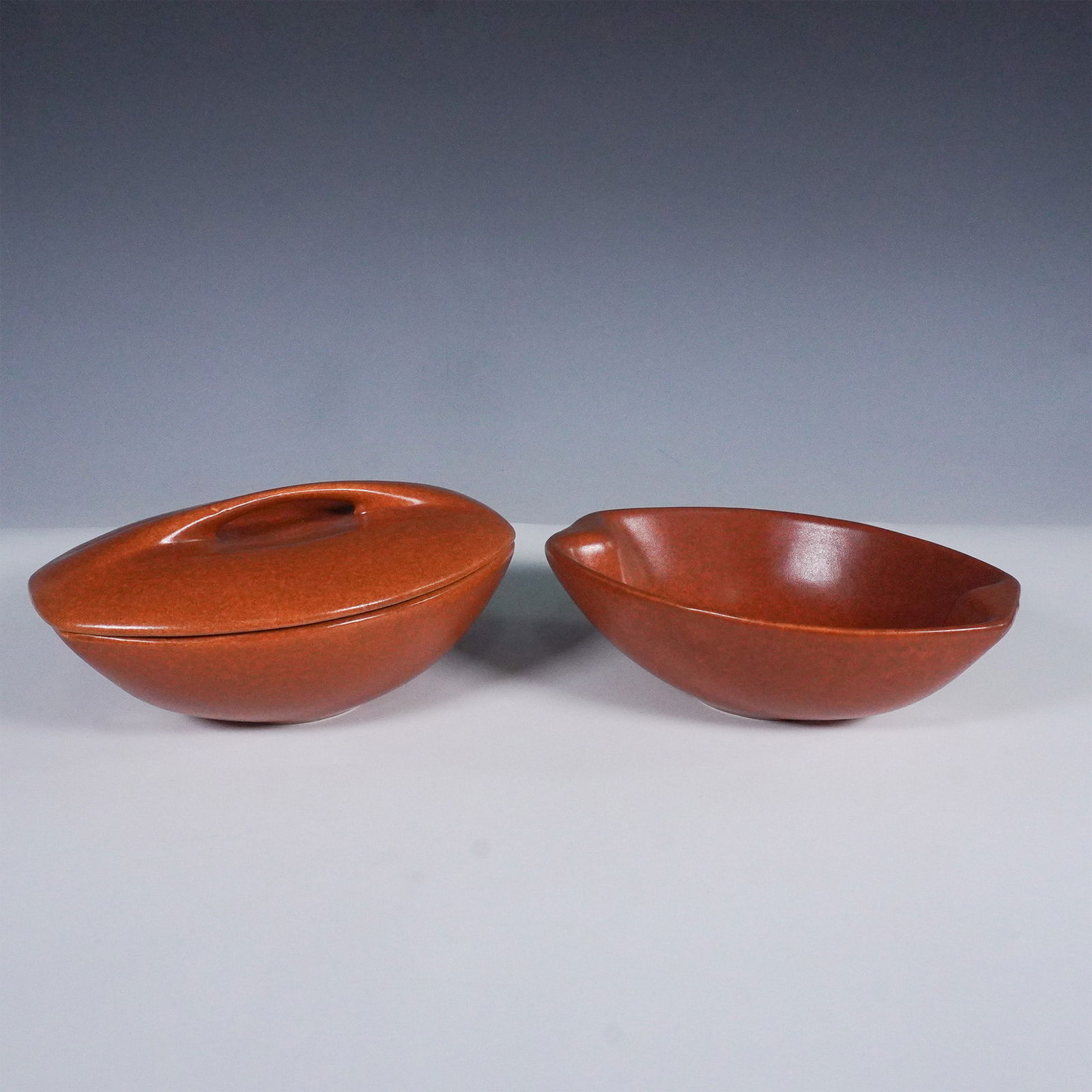 2pc Raymor by Roseville Pottery Casserole Set: Elegant mid-century modern casserole set by Roseville Pottery for the Raymor line, featuring two sleek, oval-bodied forms in a rich reddish-brown glaze. One bowl comes with its original streamlined li