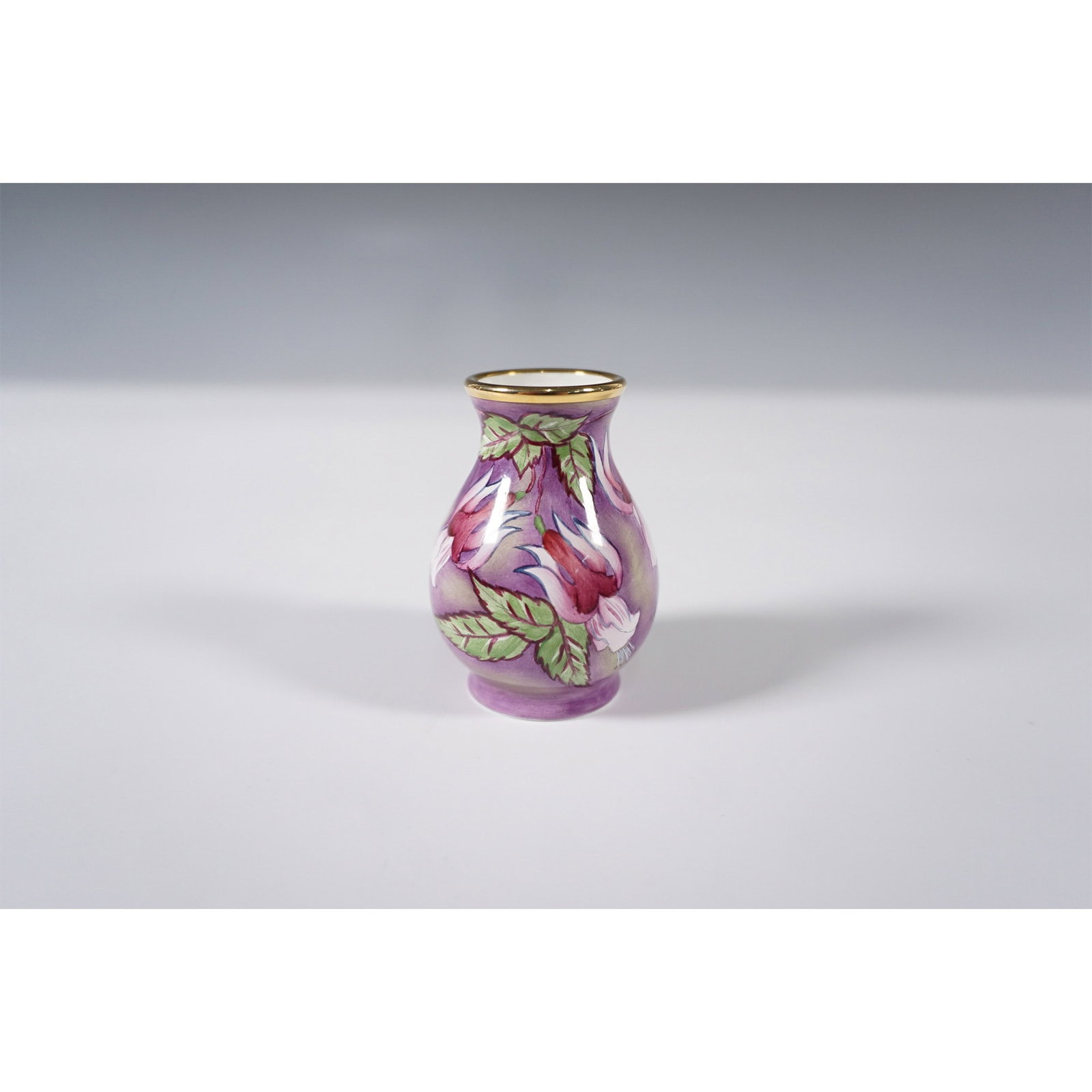 Moorcroft Enamel Fuchsia Vase: This Moorcroft enamel mini vase features a delicate hand-painted design of purple fuchsia flowers set against a soft lavender background. Accented with rich green foliage, the floral motif is rendered
