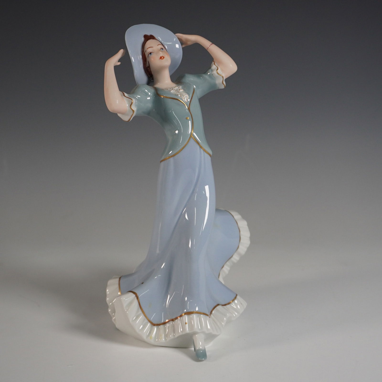 Royal Dux Bohemia Porcelain Figurine: A stunning Royal Dux Bohemia porcelain figurine featuring a gracefully posed woman in a flowing pastel blue gown with gold trim, complemented by a wide-brimmed hat. This exquisite piece highlights the