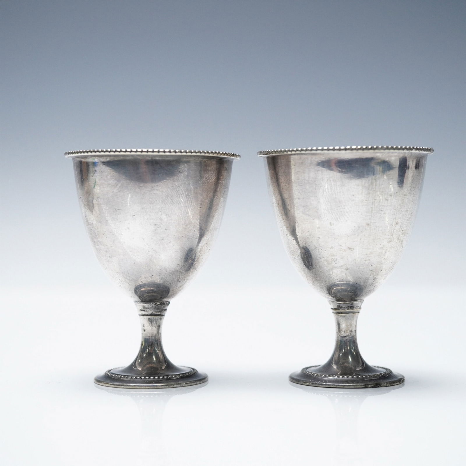 2pc Silver Plated Egg Cups: A pair of silver-plated egg cups featuring a classic beaded rim and pedestal base design. Each cup measures 2.5"H x 2" in diameter. The interiors have a subtle gold wash, adding a touch of elegance. A