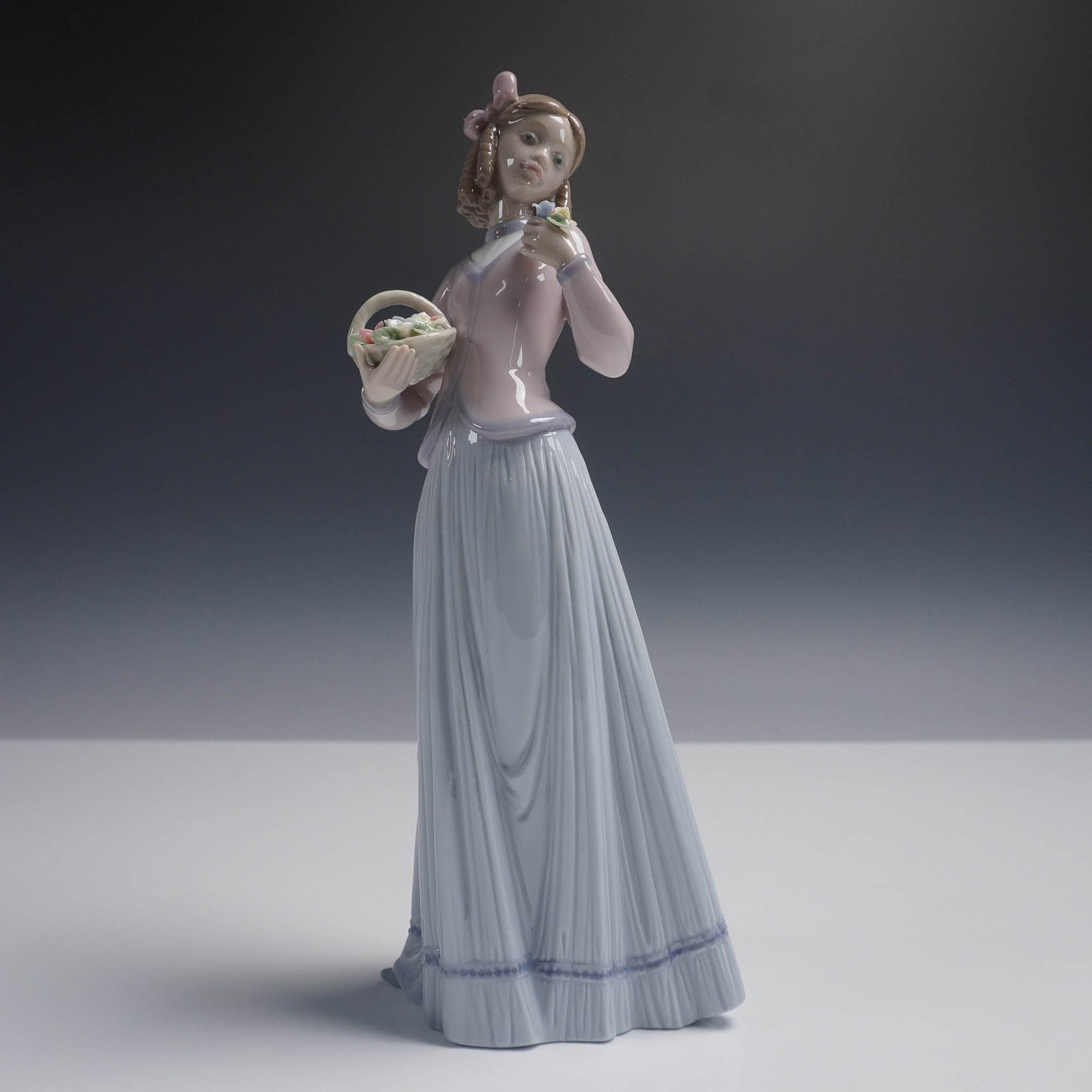 Innocence in Bloom 01007644 - Lladro Porcelain Figurine: Lladro porcelain figurine titled Innocence in Bloom (01007644), featuring a young girl in a flowing lavender and white dress holding a flower basket and admiring a delicate blossom. This elegant piece