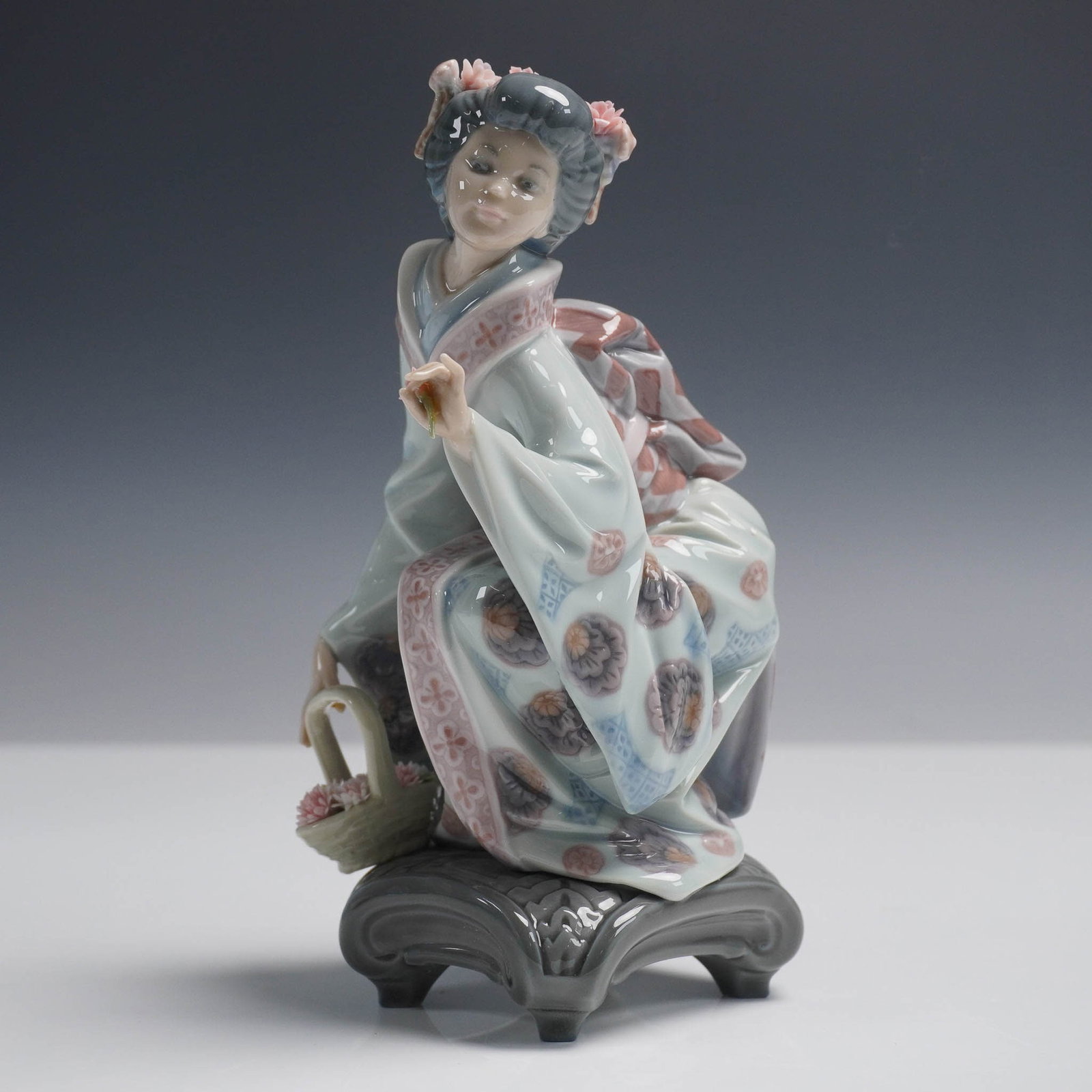 Yuki 1001448 - Lladro Porcelain Figurine: This Lladro figurine, Yuki, portrays a young Japanese woman in a detailed kimono crouched beside a flower basket. Sculpted by Salvador Deb?n, the piece features intricate pattern work, delicate flower