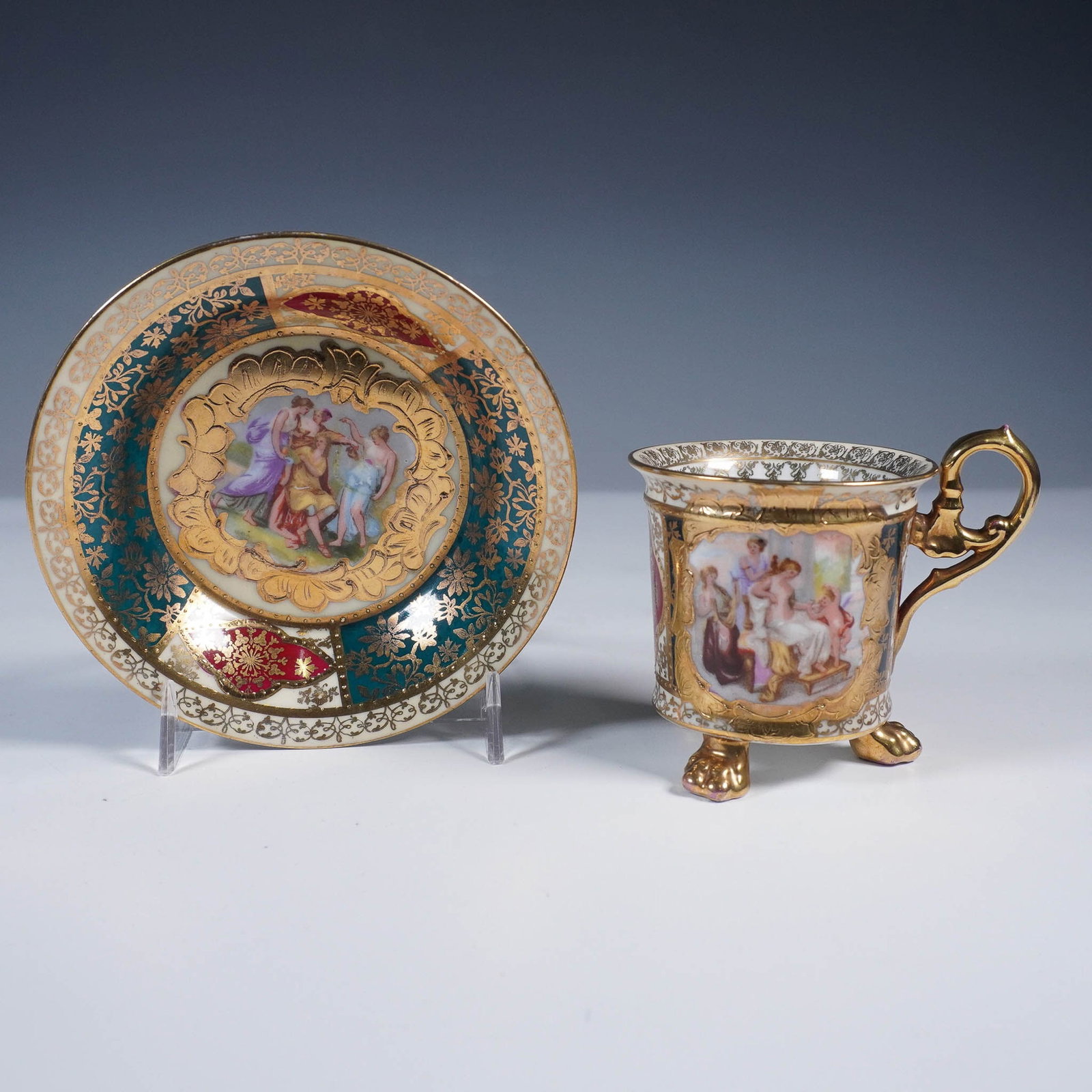 Vienna-Style Cup and Saucer, Mythological Scene: Decorative cup and saucer set in the style of Royal Vienna, featuring a scene framed in ornate gilding. The cup rests on three gilt lion paw feet and includes a scroll handle with foliate detailing. T