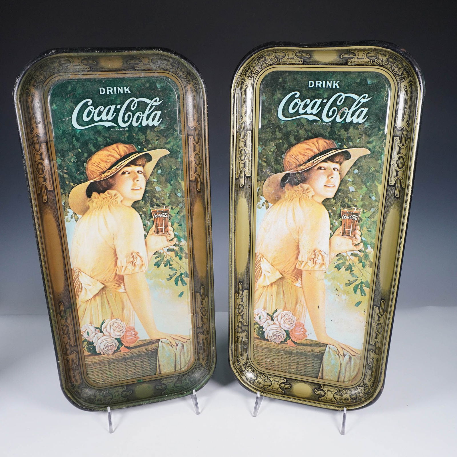 2pc Coca-Cola Girl Tin Tray Set: Pair of Coca-Cola tin trays featuring the iconic image of a young woman in a yellow dress holding a glass of Coca-Cola, set against a lush green background with roses and a wicker basket. These reprod