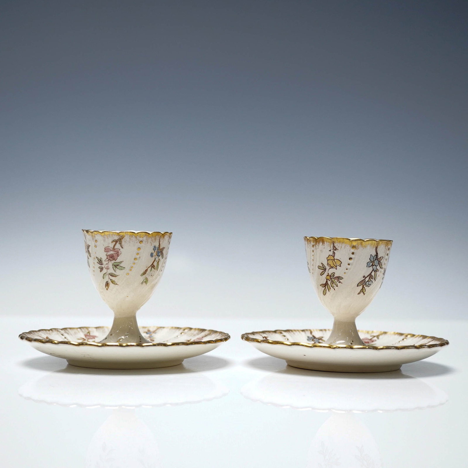 Pair Sarreguemines Louis XV Floral Egg Cups with Saucers: Pair of antique French faience egg cups with matching saucers by Sarreguemines, decorated in the Louis XV pattern. Each pedestal-form egg cup and scalloped saucer features delicate hand-painted floral