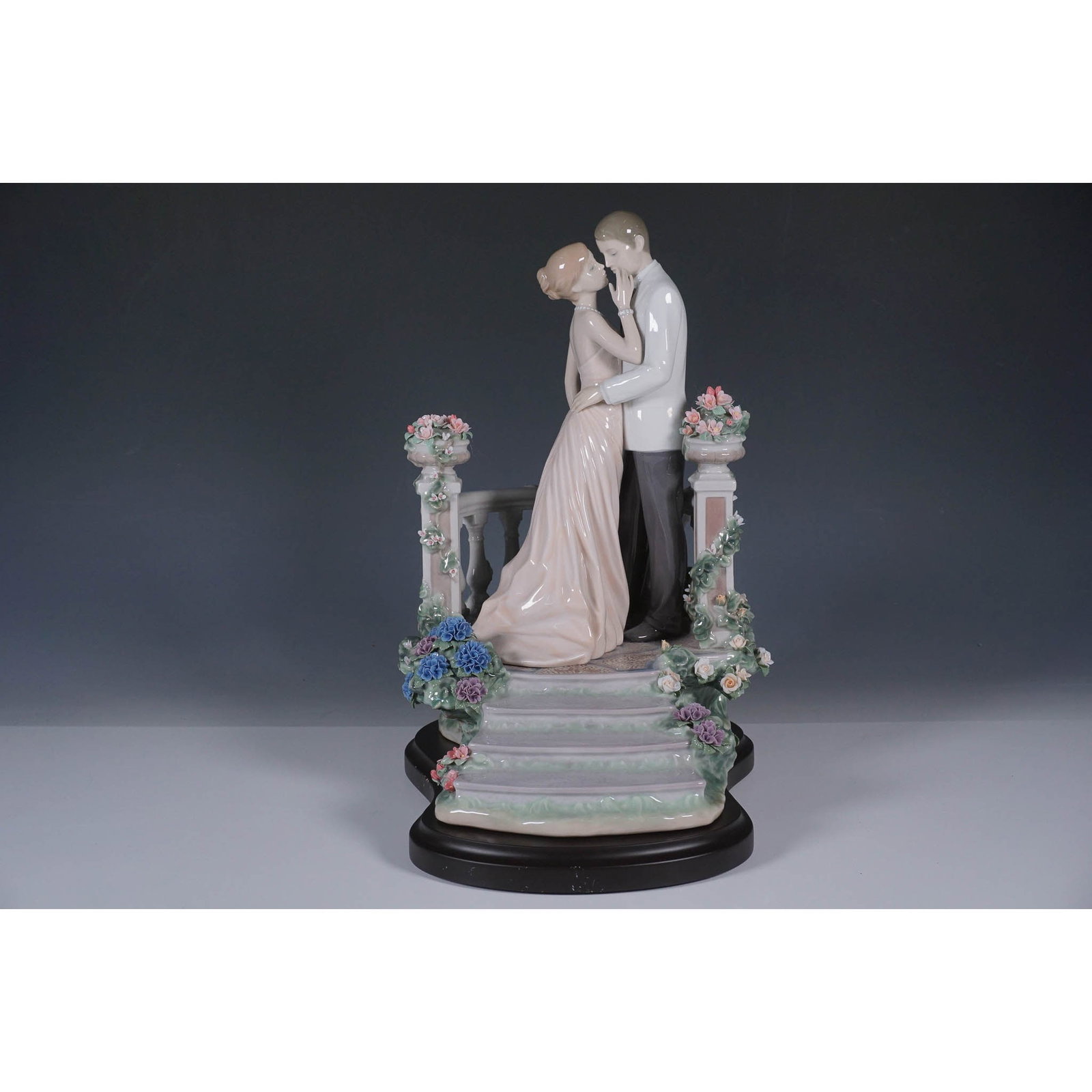 Moonlight Love 1007695 - Lladro Figurine with Base: This is an exquisite Lladro porcelain figurine, a masterful depiction of romantic elegance and detailed artistry. The scene features a couple tenderly embracing on a flower-adorned staircase, framed b