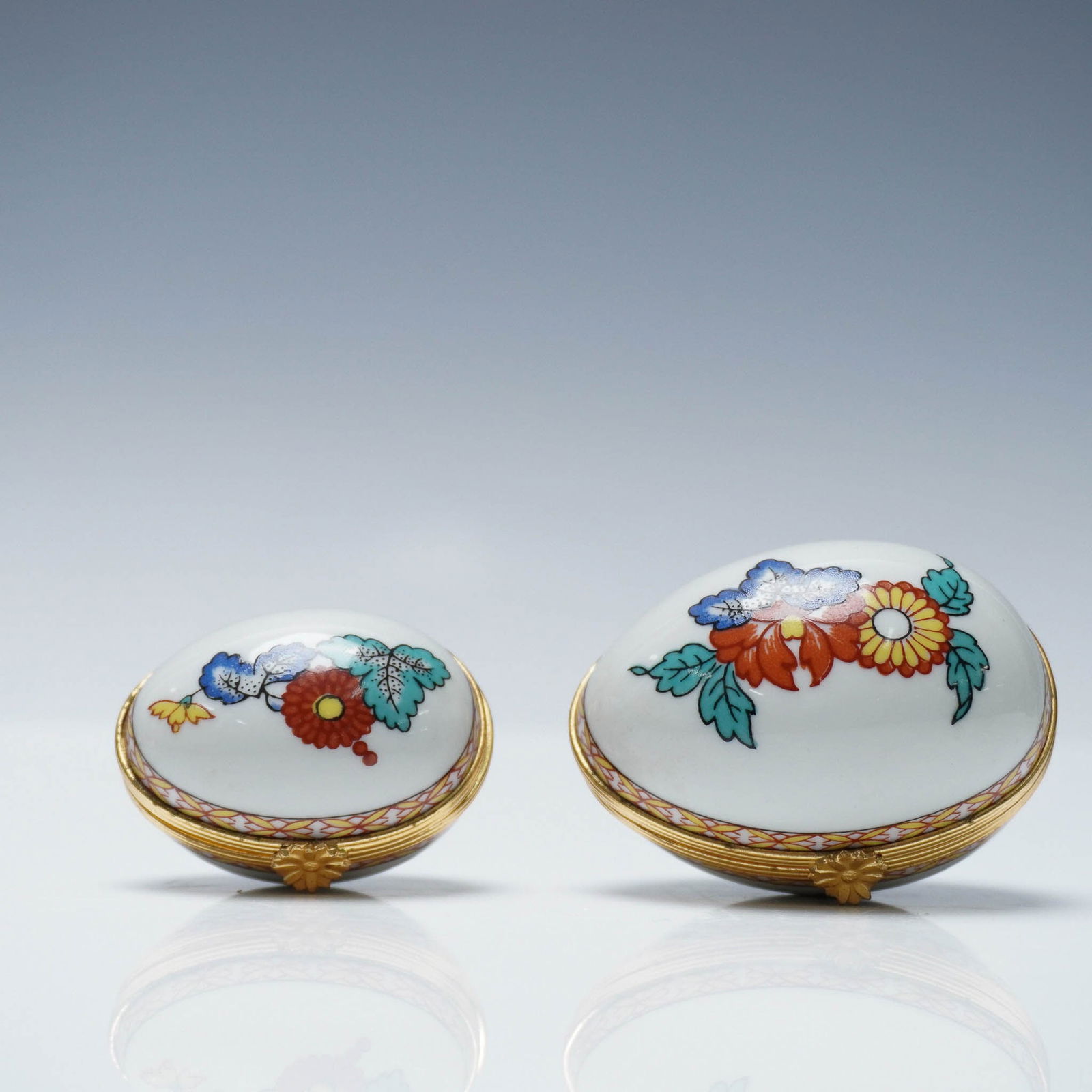2pc Limoges Castel Floral Egg Treasure Boxes: Pair of Limoges Castel trinket boxes from France, each crafted in an egg shape with hand-painted floral motifs in vibrant tones of red, yellow, blue, and green. Both boxes feature finely detailed gilt