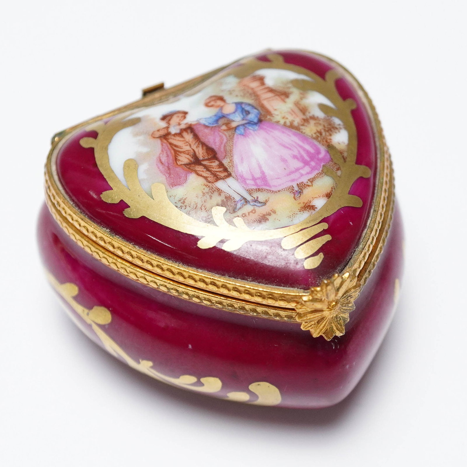 Limoges France Porcelain Heart Treasure Box: Hand-painted Limoges porcelain trinket box in a heart shape, featuring a romantic 18th-century courting scene on the lid surrounded by intricate gilt detailing. The box is a deep cranberry red with go