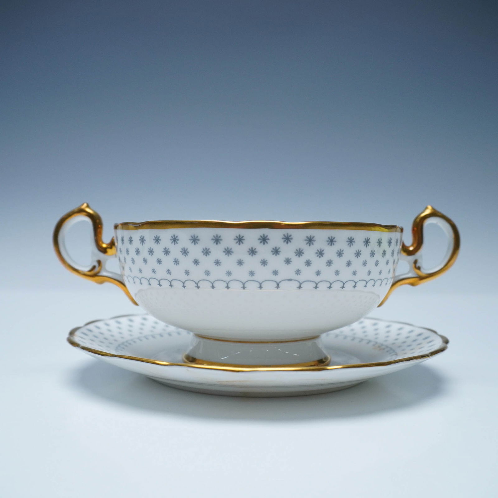 2pc Royal Chelsea English Bone China Gray & Gold Soup Cup and Saucer: A refined Royal Chelsea English bone china cream soup cup and saucer set, featuring an intricate blue starburst pattern accented with rich gold trim. The elegantly shaped cup showcases ornate double h