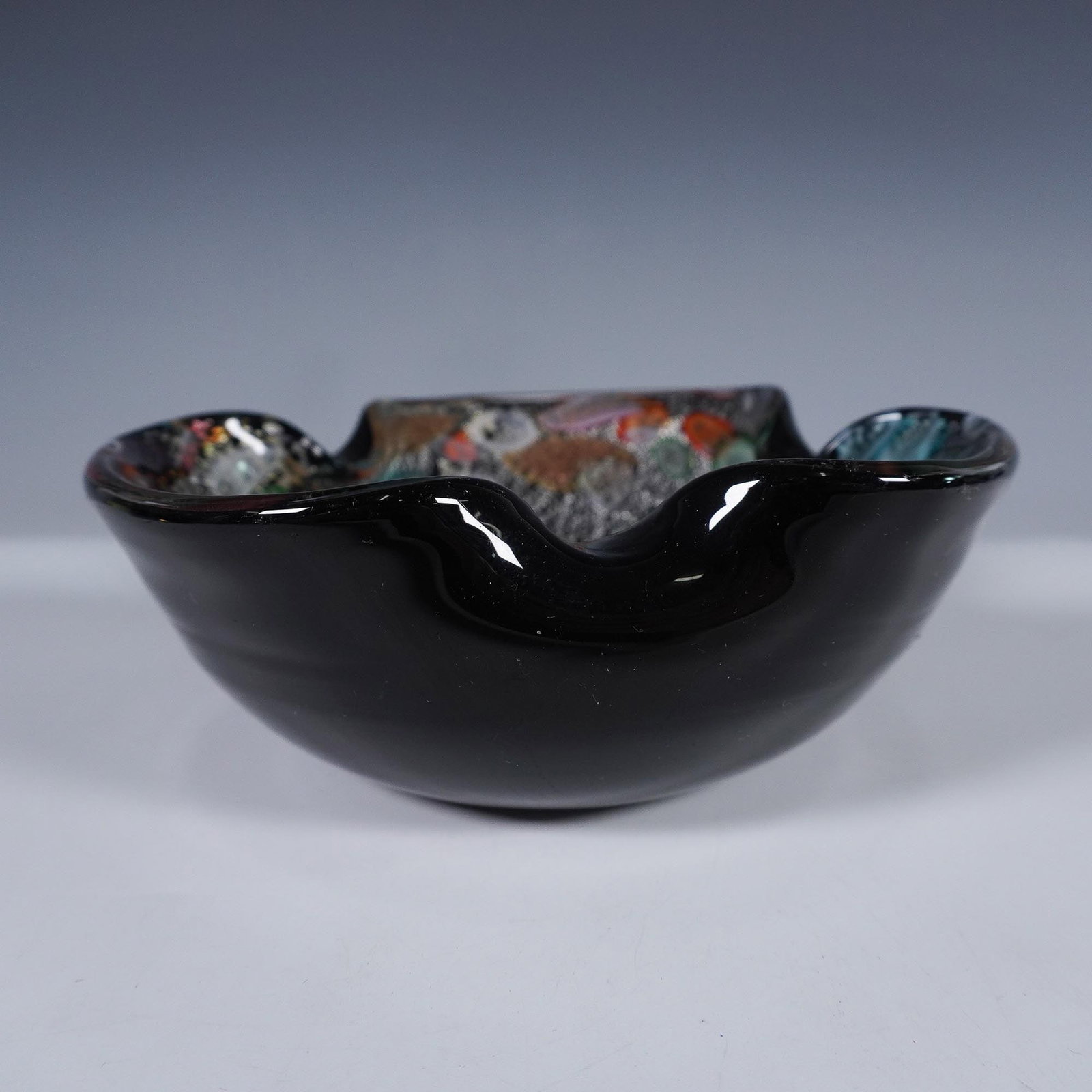Murano Style Abstract Art Glass Bowl: Murano styled cased black glass bowl with multi-color iridescent design with rolled edges. Issued: 21st century Dimensions: 2.5"H x 7" dia.
