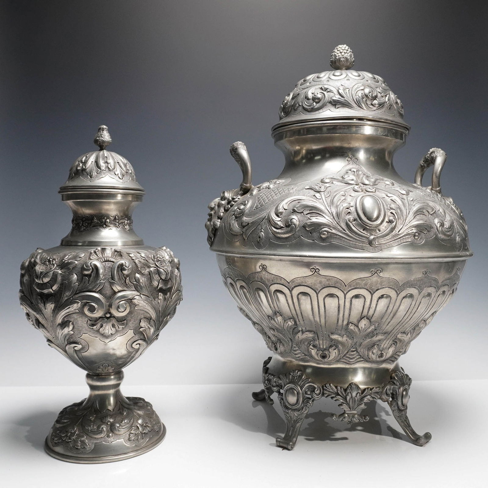 Pair of J. Dores Ornate Portuguese Pewter Lidded Wine Urns: A highly decorative pair of Portuguese pewter vessels richly adorned in high-relief Rococo inspired ornamentation. Each vessel features scrolling acanthus motifs, cartouches, and stylized floral accen