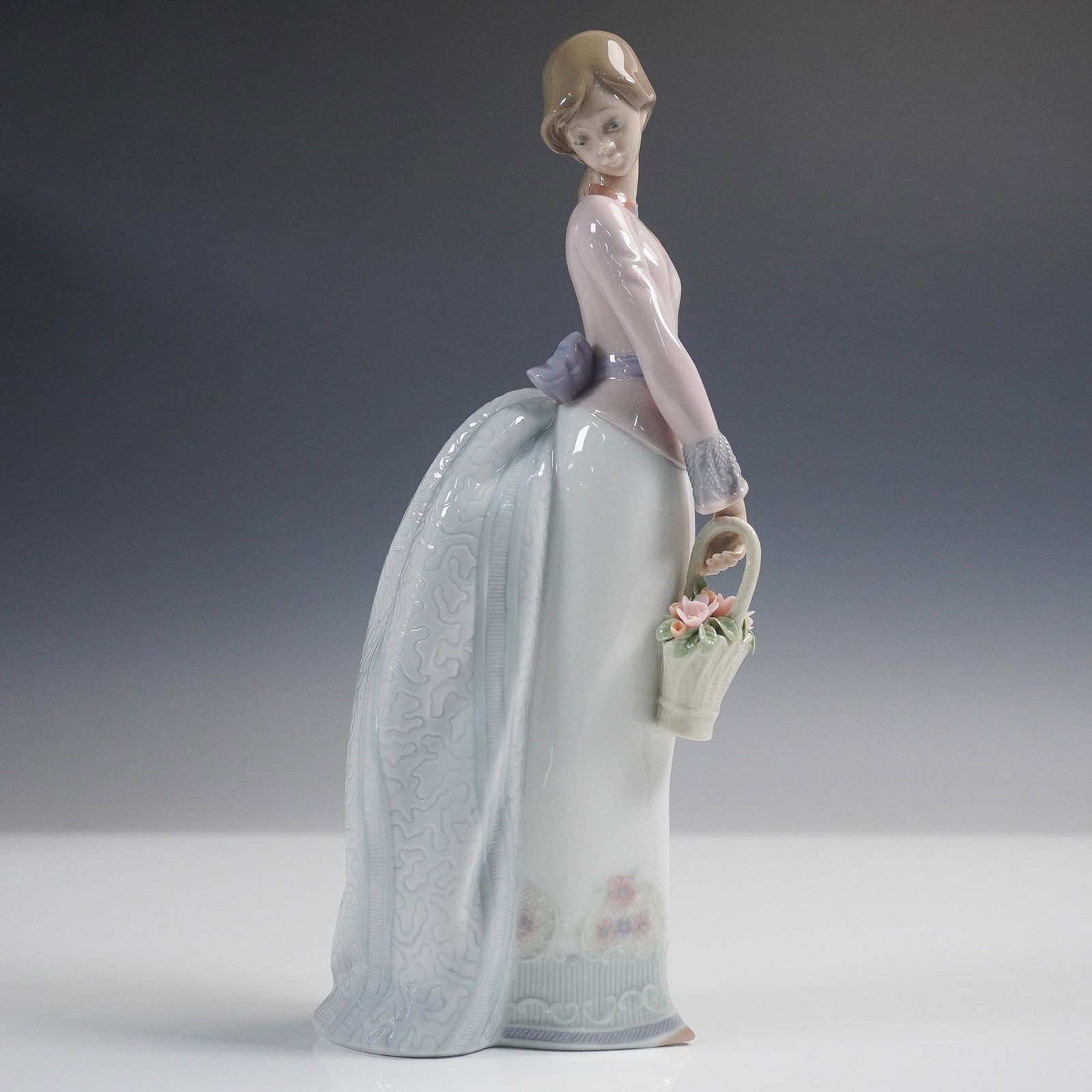 Lladro Collectors Society Porcelain Figurine, Basket of Love 1007622: This glazed porcelain piece depicts a young woman in a long white and lavender gown, carrying a basket of pastel-toned flowers. The dress features a textured train and ribbon detail at the back, and f