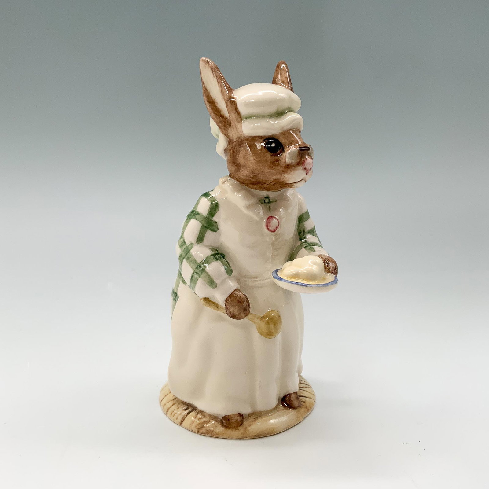 Royal Doulton Bunnykins Figurine, Cook DB85: Royal Doulton Bunnykins figurine titled Cook, DB85. This delightful figurine showcases a bunny chef dressed in a green and white plaid apron, holding a spoon and a dish. Backstamp indicates production