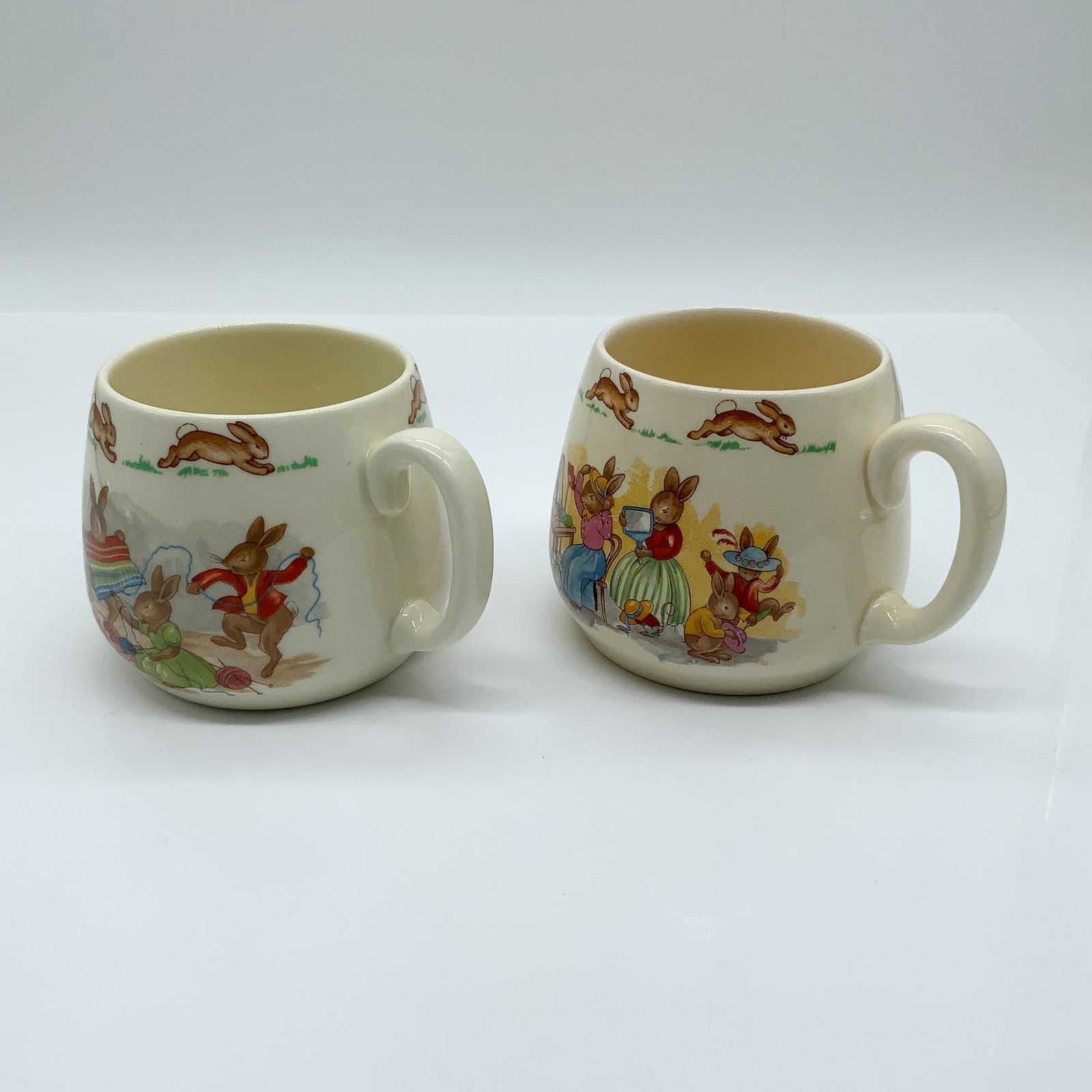 2pc Royal Doulton Bunnykins Mugs, Childrens Cups: his lot features a delightful pair of Royal Doulton Bunnykins mugs, each showcasing charming hand-painted scenes of playful rabbits in a variety of activities. These mugs, part of the Bunnykins series