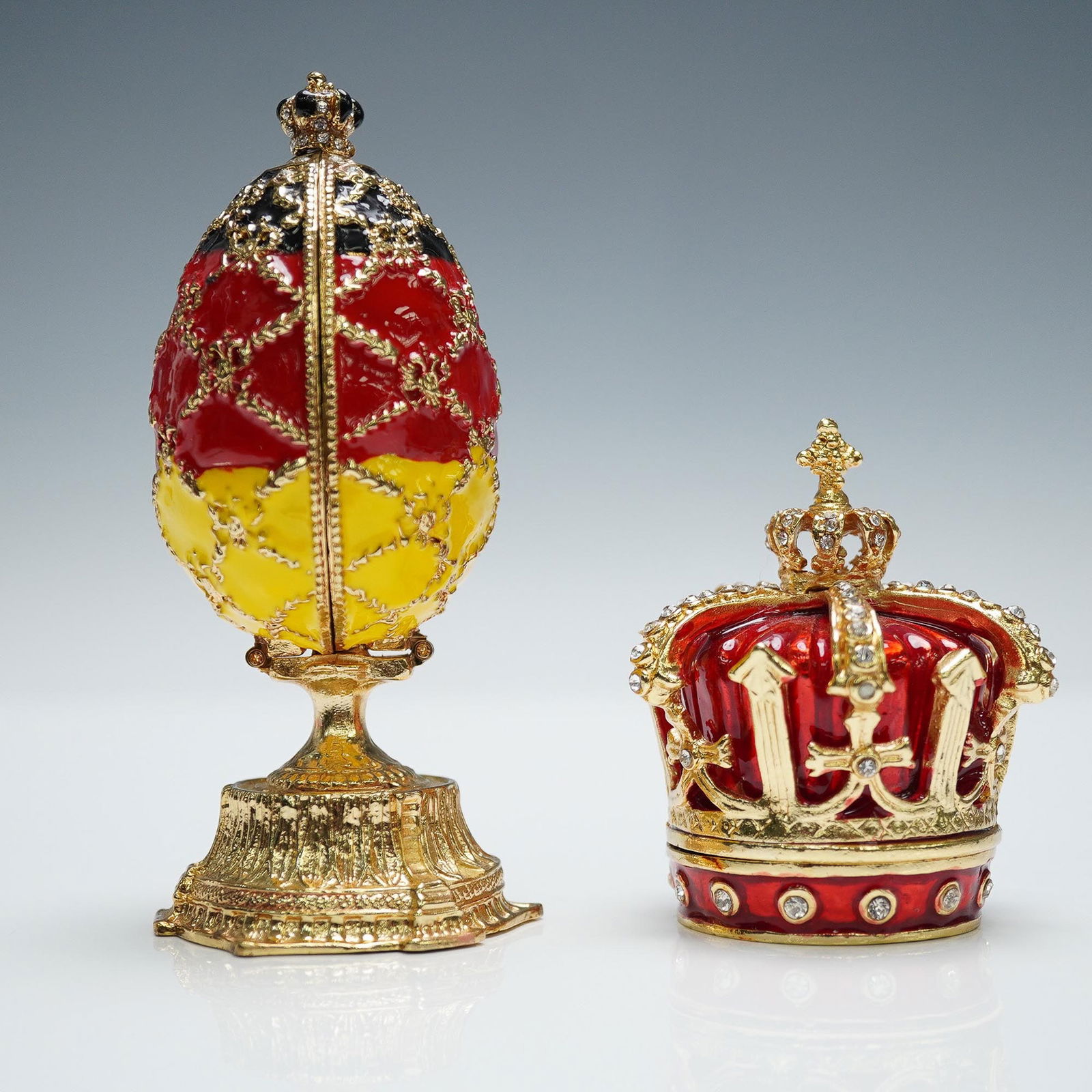 2pc QIFU Jeweled Enamel Boxes, World Cup Egg and Crown: Set of two decorative enameled decorative eggs, including a crown shaped box with red enamel and rhinestone accents, and an egg shaped box adorned in red, yellow, and black enamel with gold-tone trim