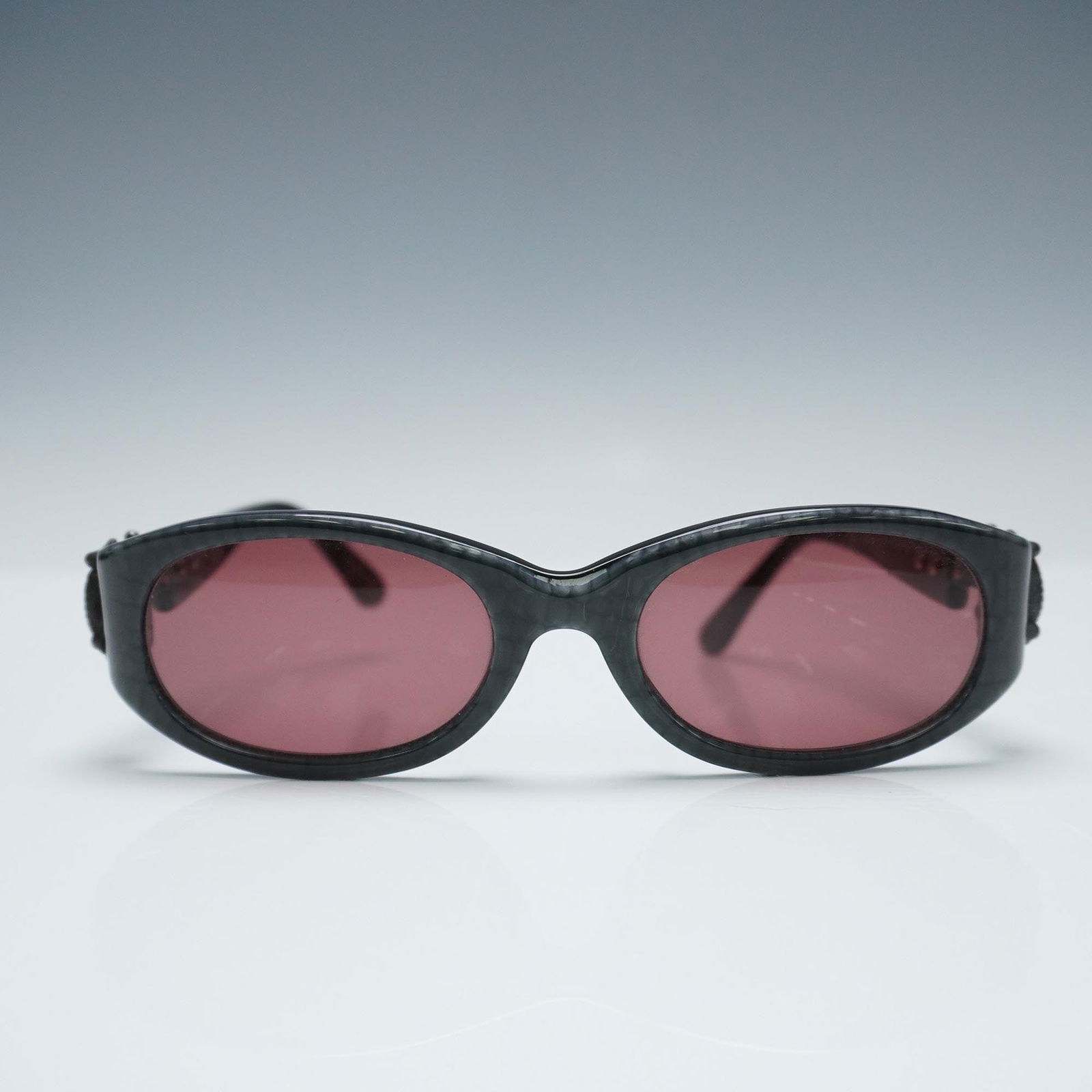 Kieselstein-Cord Surrender Sunglasses with Alligator Hinge: Kieselstein-Cord designer sunglasses featuring a bold oval frame in a dark textured finish with rose-tinted lenses. This Surrender model is crafted in Japan and showcases the brand's iconic sculptural