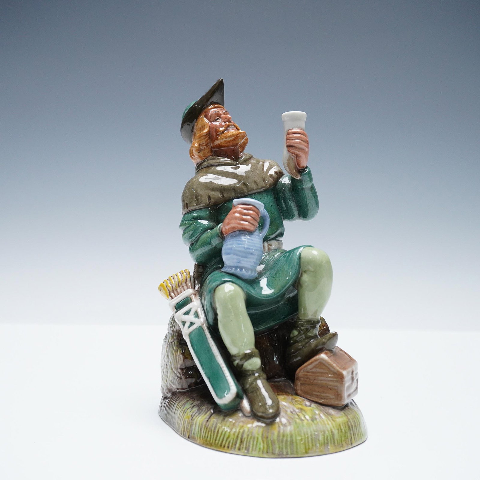 Robin Hood HN2773 - Royal Doulton Figurine: Royal Doulton figurine titled Robin Hood HN2773, depicting the legendary English folk hero in traditional green attire, seated with a goblet raised and a jug in hand. Sculpted by Mary Nicholl, this pi