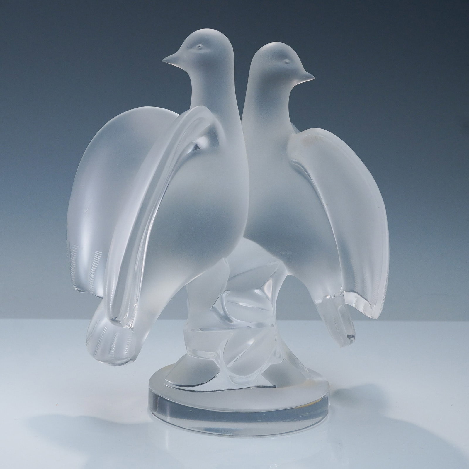 Lalique Crystal Figurine, Ariane Doves: An elegant frosted and clear sculpture depicting a pair of doves perched side by side with wings partially lifted in a graceful and symmetrical composition. Executed in the stunning frosted look, fine