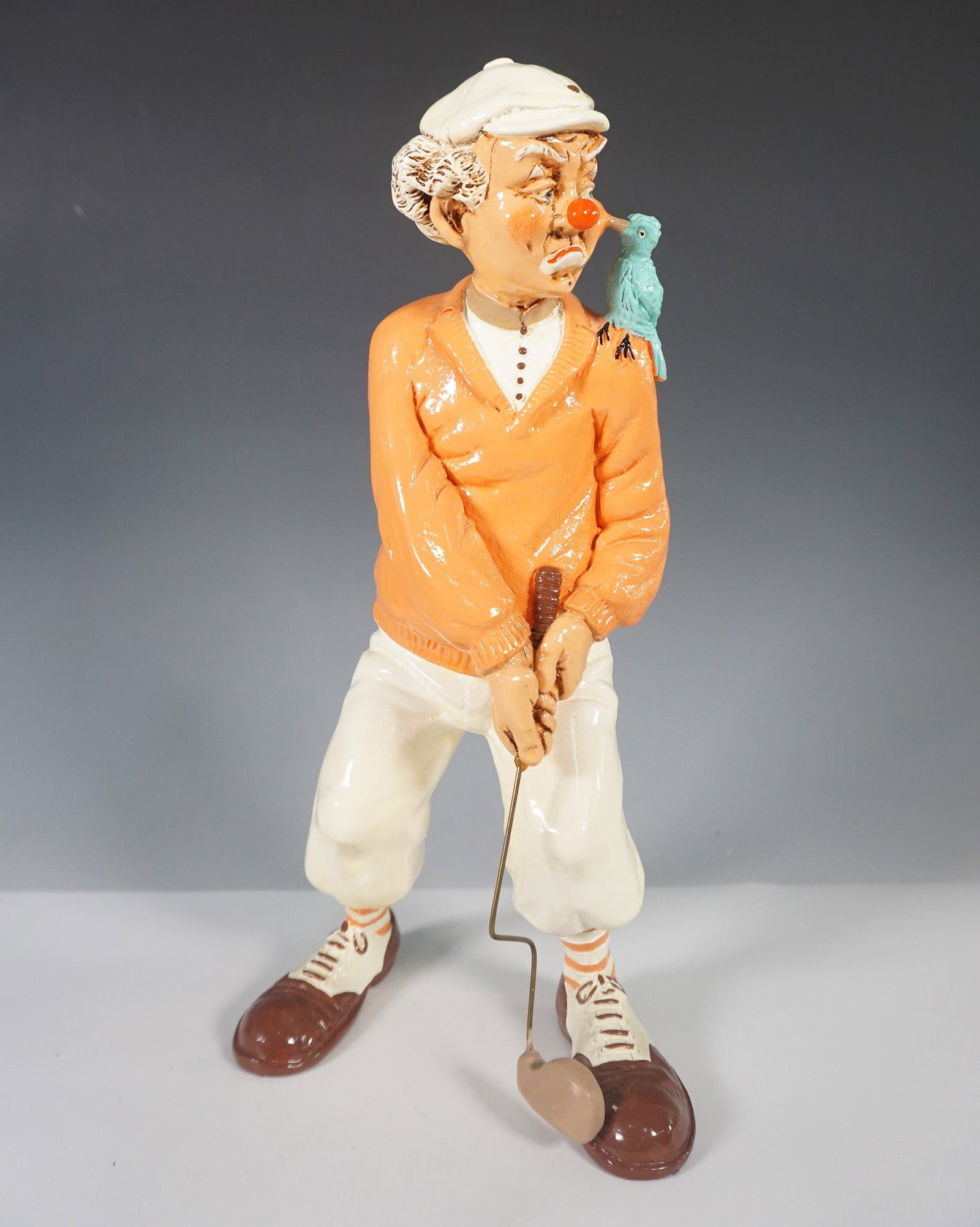 Judy's Pastime Golfer Clown Figurine: A whimsical ceramic figurine marked Judy's Past, dated 1985, depicting a comical golfer clown mid-swing. The figure features a bright orange sweater, white knickers, striped socks, and brown and white
