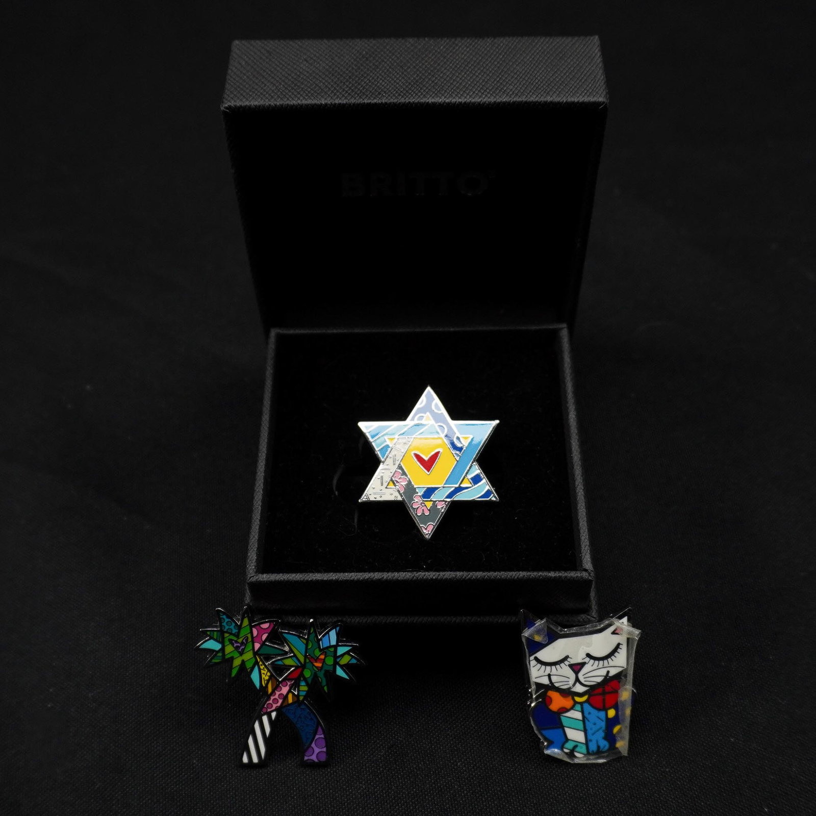 3pc Britto Enamel Pins, Star of David, Cat & Palm Trees in Britto Box: Three-piece set of Romero Britto enamel pins, each featuring the artist's signature pop art style and marked with the Britto backstamp. The collection includes a vibrant Star of David with a central r