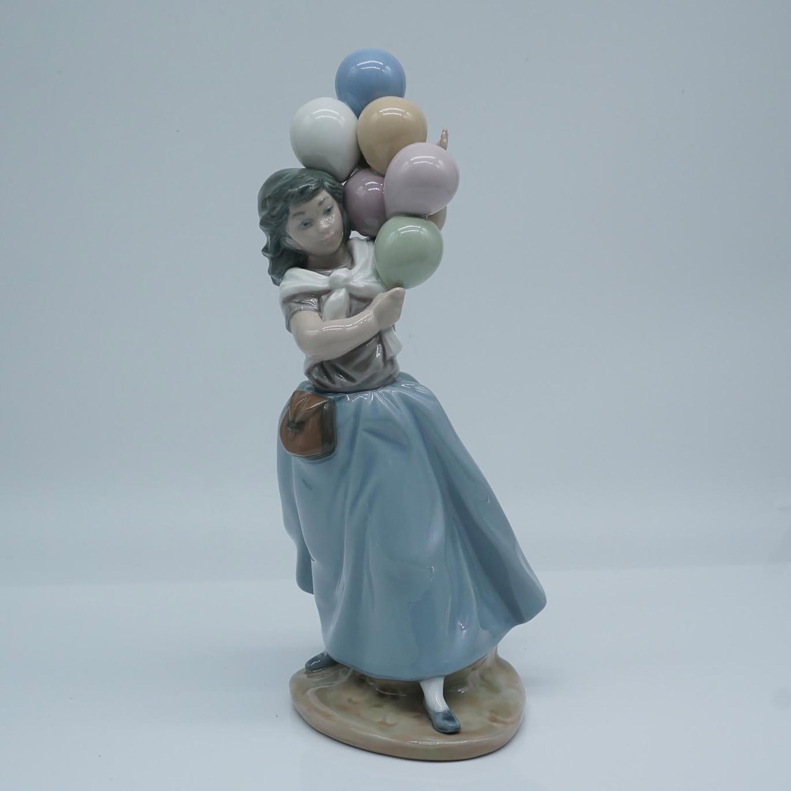 Lladro Porcelain Figurine, Balloons For Sale 1005141: A glazed finish figure depicting a young girl carring a bouquet of colored balloons in soft pink, cream, and green hues. She wears a flowing light blue skirt with a brown pouch and her gentle expressi