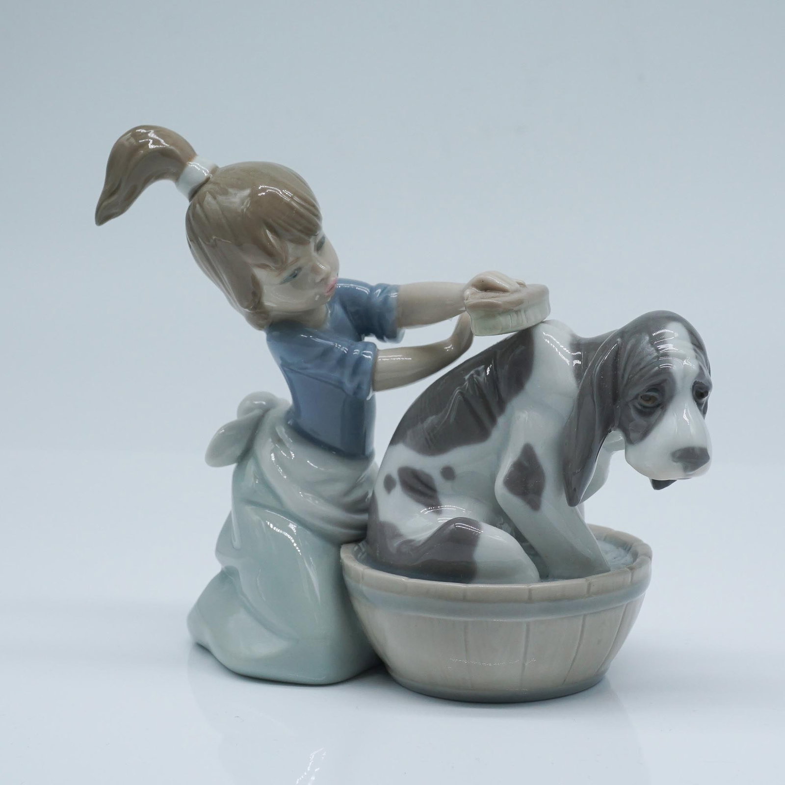 Lladro Porcelain Figurine, Bashful Bather 1005455: This glossy porcelain figurine titled Bashful Bather, depicts a young girl attentively washing her floppy eared dog, who sits patiently in a tub. The composition captures a tender moment, highlighting