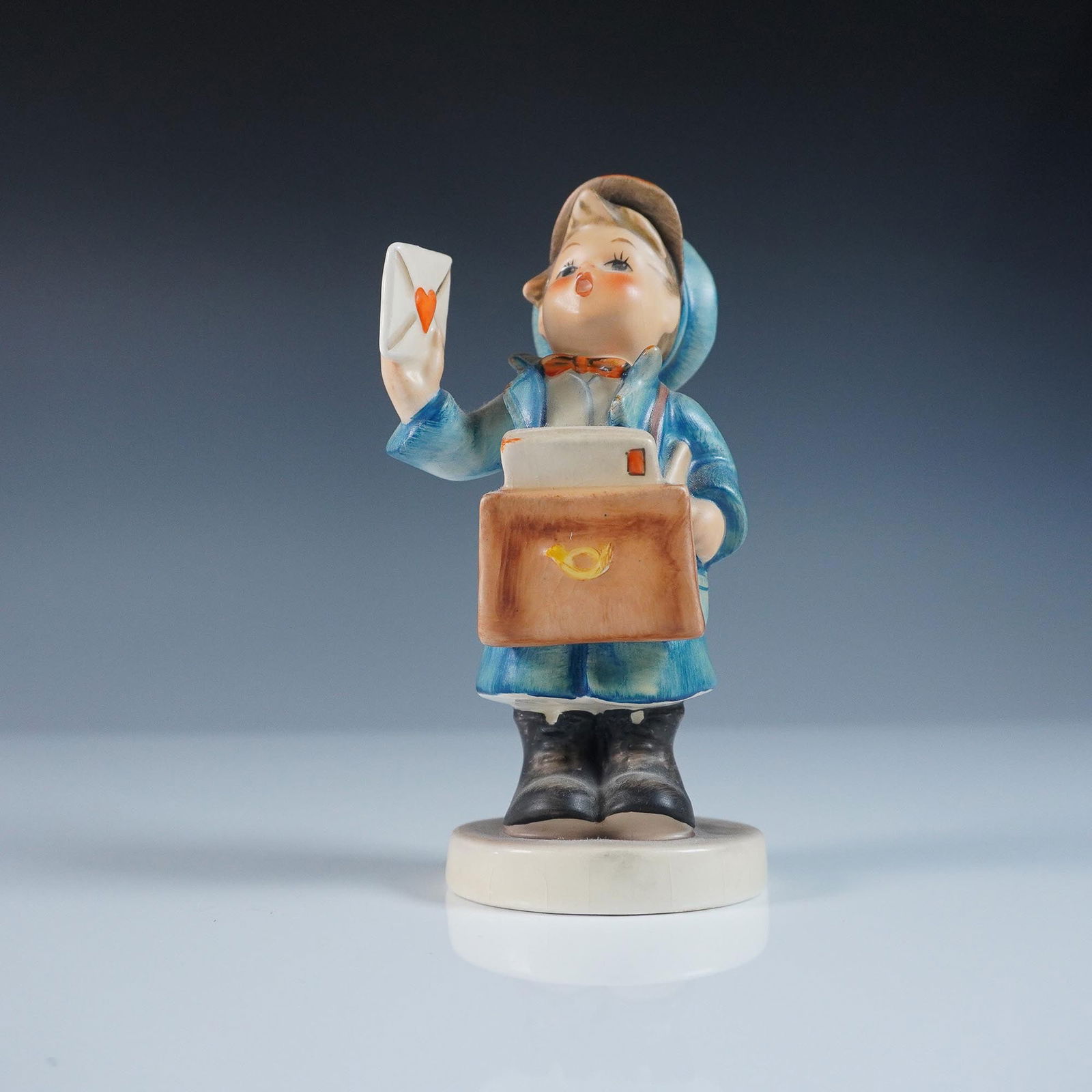 Goebel Hummel Figurine - Mailman: Goebel Hummel figurine titled Mailman, featuring a young postal carrier dressed in a blue coat and hat, holding a letter with a red heart. With a satchel marked by a postal horn this charming piece em
