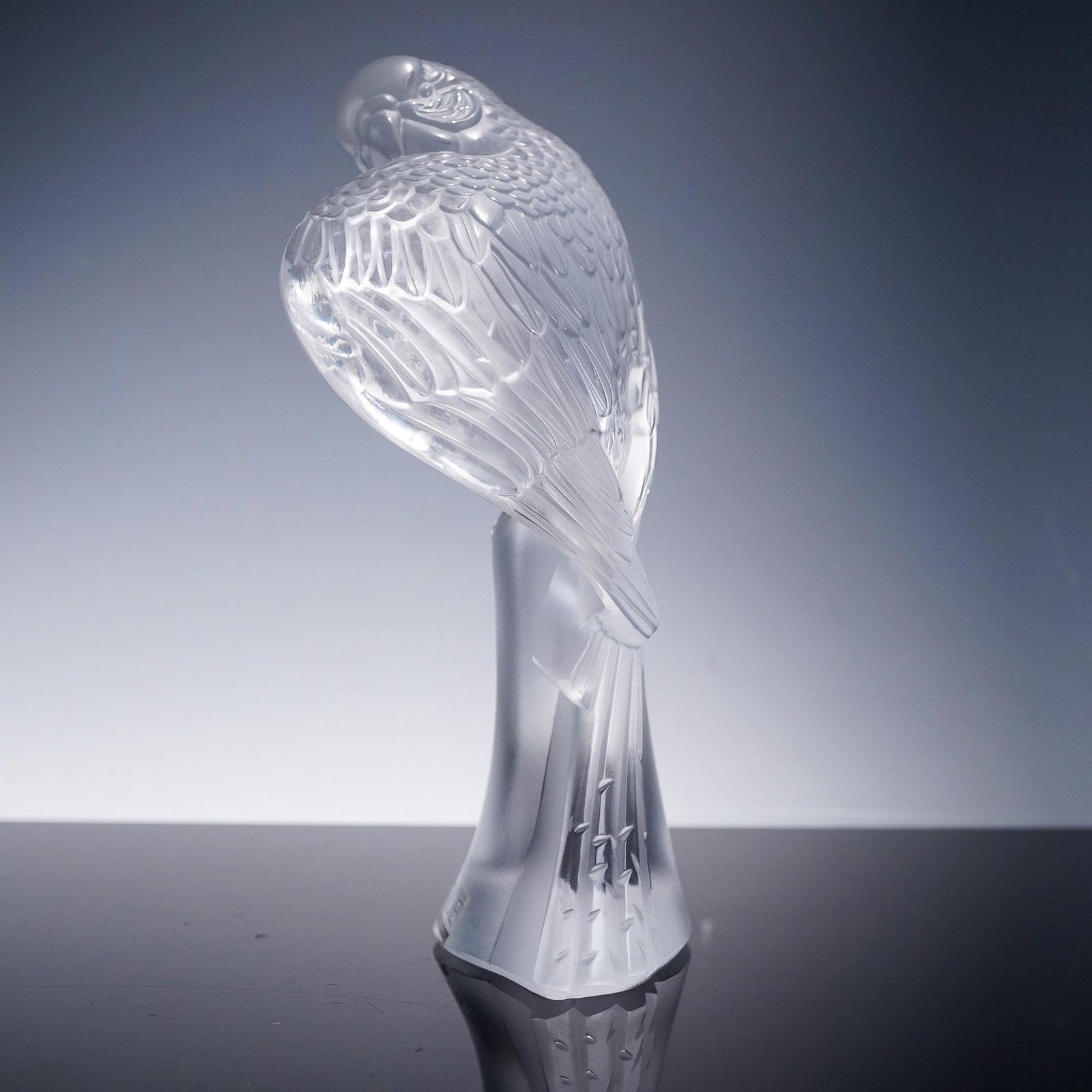 Lalique Clear Crystal Figurine, Parrot: A striking Lalique clear crystal figurine depicting a majestic parrot perched gracefully, with finely detailed feathers and a sleek, curved form. The frosted and polished surfaces create a beautiful c
