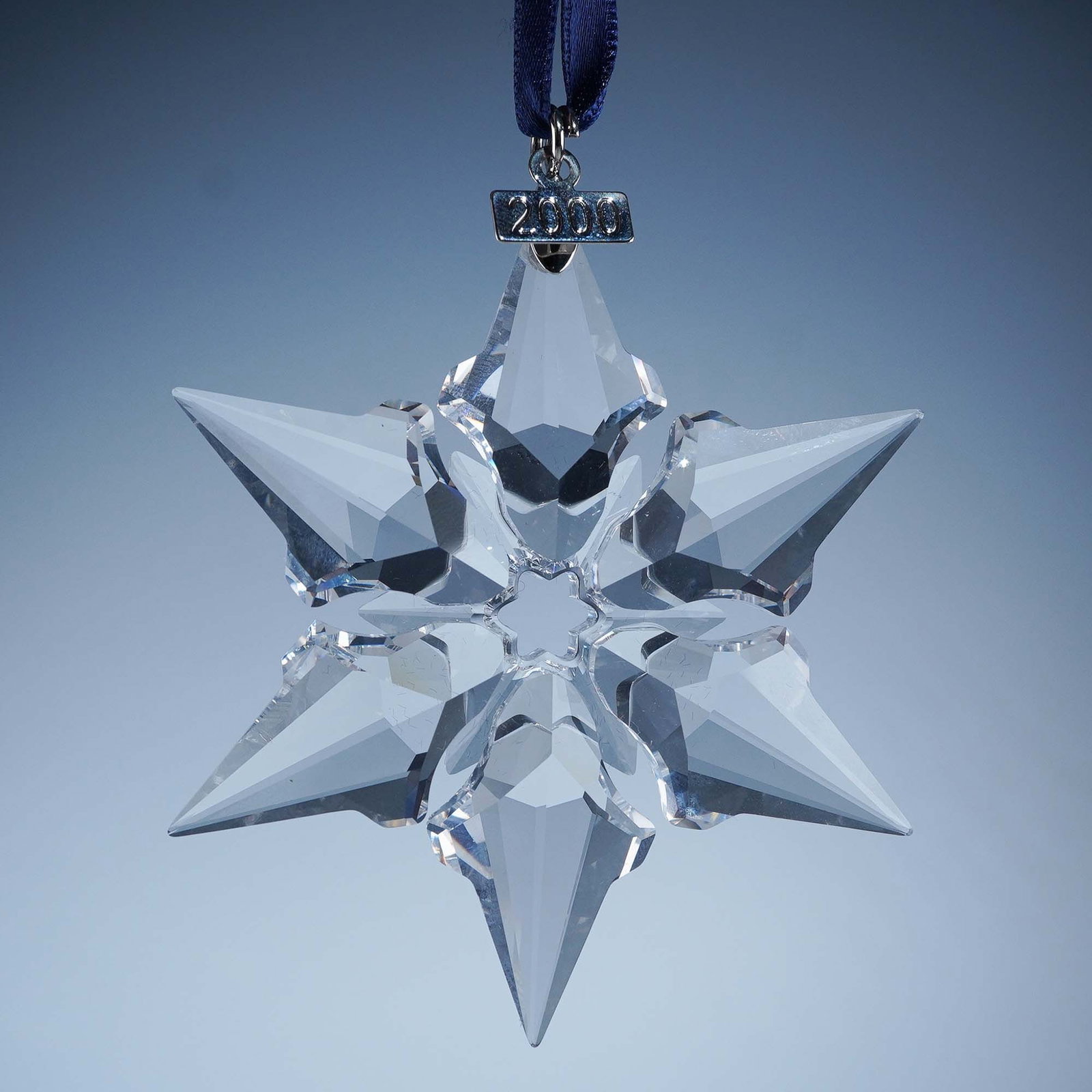 Swarovski Crystal 2000 Christmas Ornament: Featuring a six-point star design with faceted surfaces arranged around a small central opening. Each point is elongated with angular edges, forming a symmetrical snowflake pattern. The ornament is su