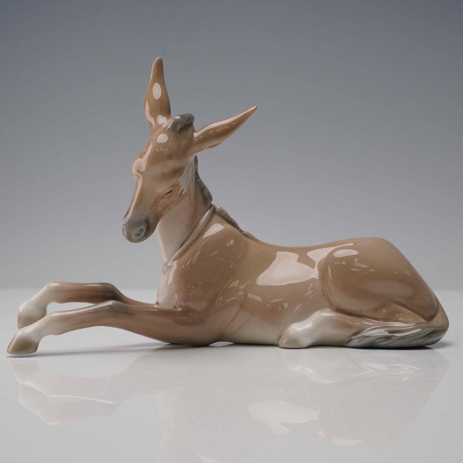 Lladro Porcelain Figurine, Donkey 1004679: Lladro porcelain figurine titled "Donkey" (model 1004679), depicting a serene, reclining donkey in soft brown and cream hues with a high-gloss finish. This elegant sculpture captures the animal's gent