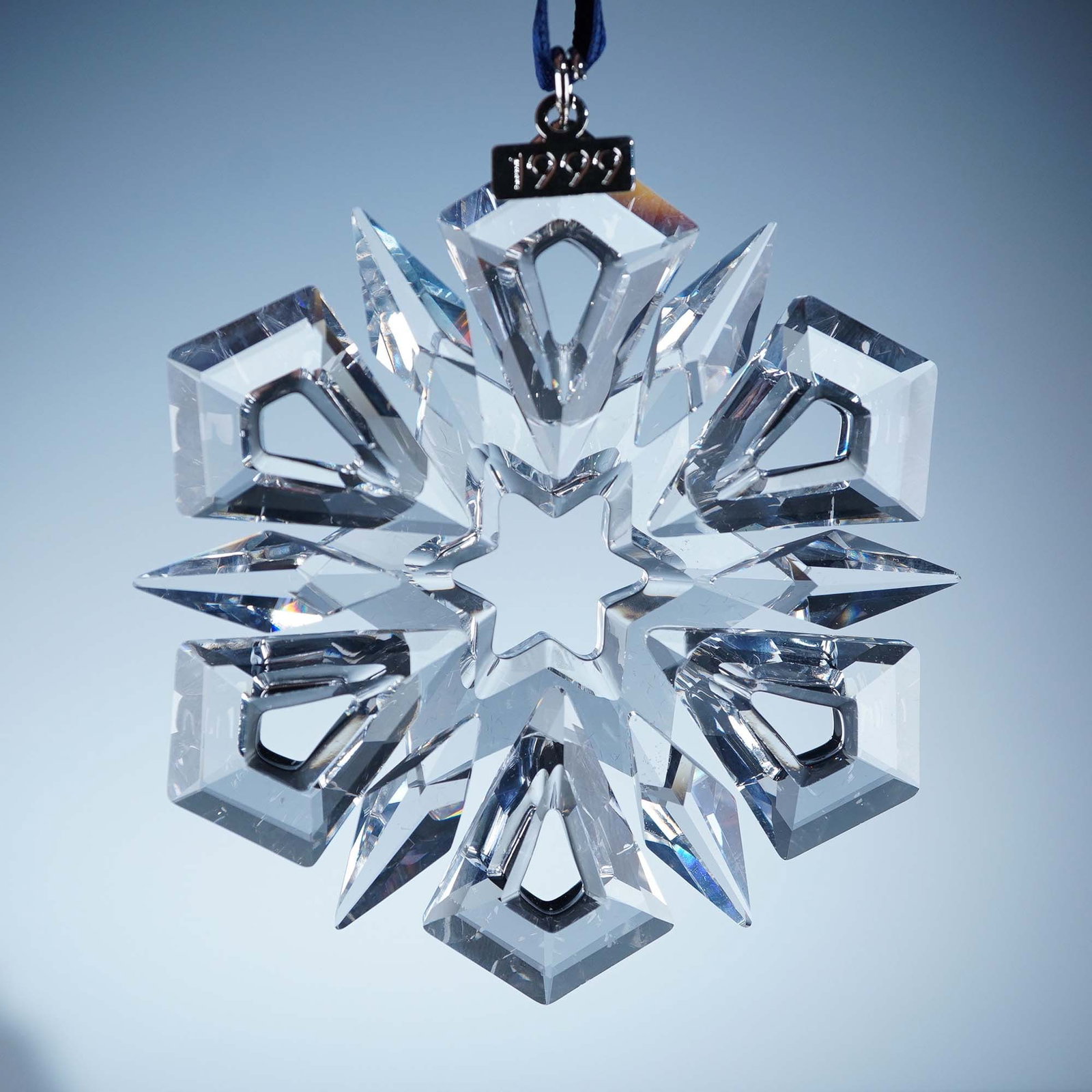 Swarovski Crystal 1999 Christmas Ornament: Featuring a 12-point symmetrical snowflake design with alternating kite-shaped and pointed crystal arms radiating from a central star-shaped cutout. The ornament includes a metal tag engraved with the