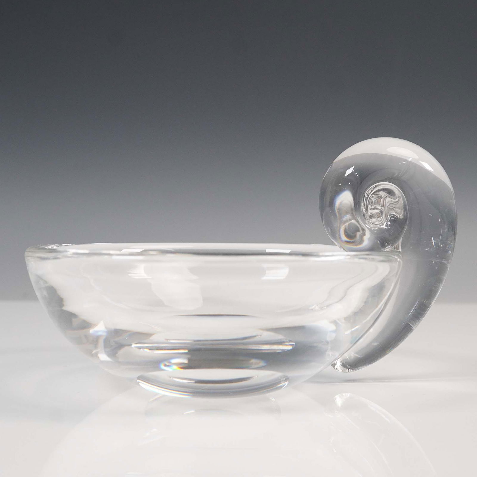 Steuben Glass Round Snail Bowl with Cloth Pouch: A clear, round bowl featuring a distinctive applied snail-form handle. The bowl is crafted with smooth, rounded walls and a flat base, while the handle rises in a coiled spiral with an internal air tr