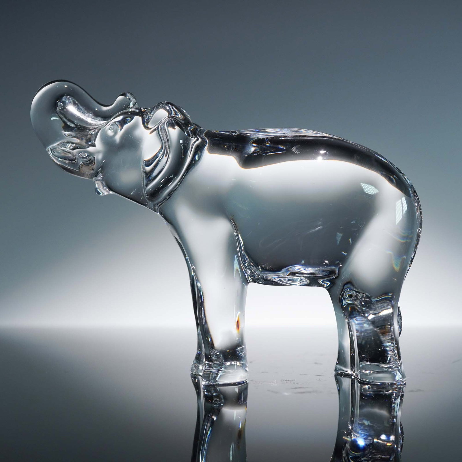 Baccarat Crystal Figurine, Elephant with Raised Trunk: This charming Baccarat crystal figurine features a stylized elephant standing in a proud pose with its trunk gracefully raised. Expertly crafted in brilliant French crystal, the form captures gentle c