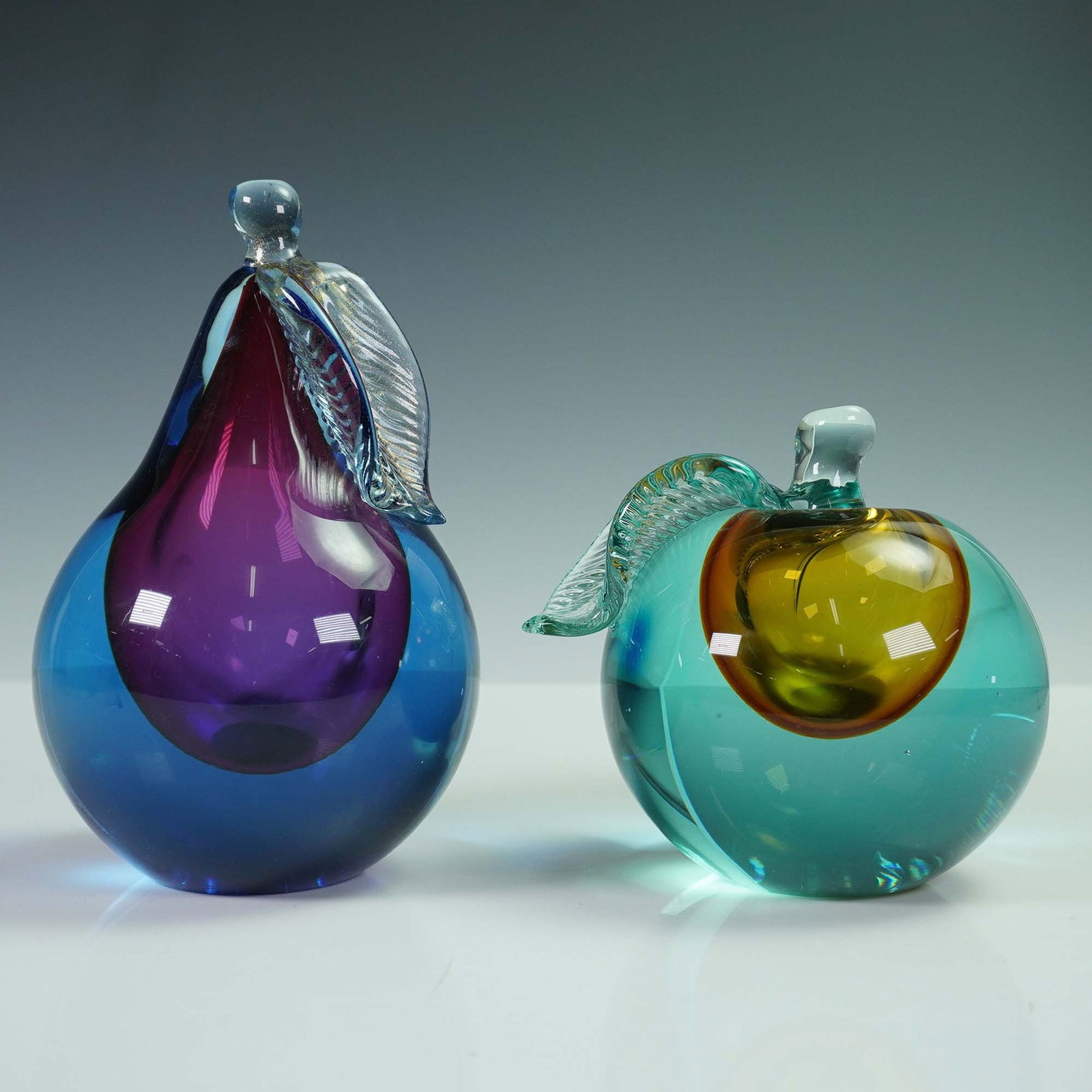 2pc Murano Somerso Fruit Figures, Pear and Apple: Pair of contemporary studio art glass fruit sculptures of a pear and apple form. Each features vibrant color layering called the Somerso technique. One with rich purple and blue hues and other with tu