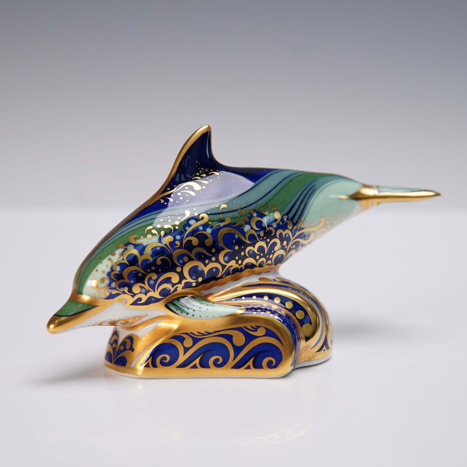 Royal Crown Derby Porcelain Paperweight, Baby Bottlenose Dolphin: This graceful Royal Crown Derby porcelain paperweight features the Baby Bottlenose Dolphin, captured mid-leap atop stylized waves. The design is adorned in vivid blue, green, and lavender tones with f
