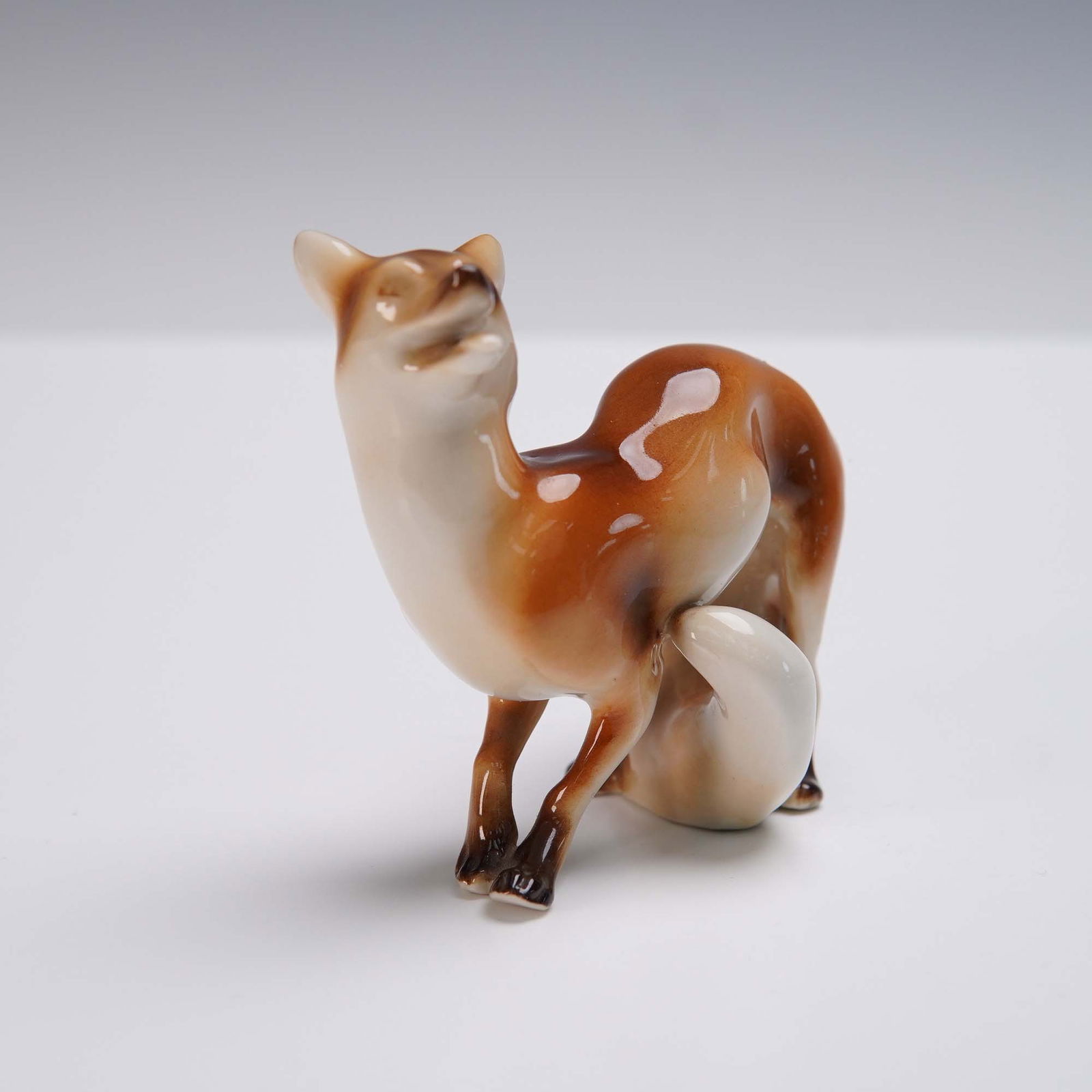 Royal Dux Porcelain Figurine, Fox: Crafted by the esteemed Royal Dux porcelain factory, this hand-painted porcelain fox figurine features a striking blend of light caramel and white glaze with finely modeled detailing in the fur and po