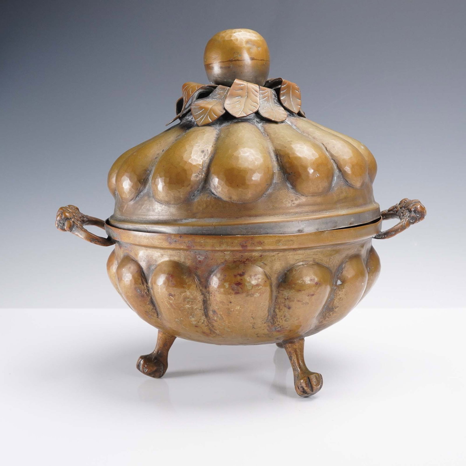Brass Hand Hammered Lidded Dish with Leaf Finial: This hand hammered brass dish features a bulbous, lobed body raised on three paw feet with scrolled floral handles. The lid is topped with a fruit-shaped finial encircled by applied figural leaves, sh