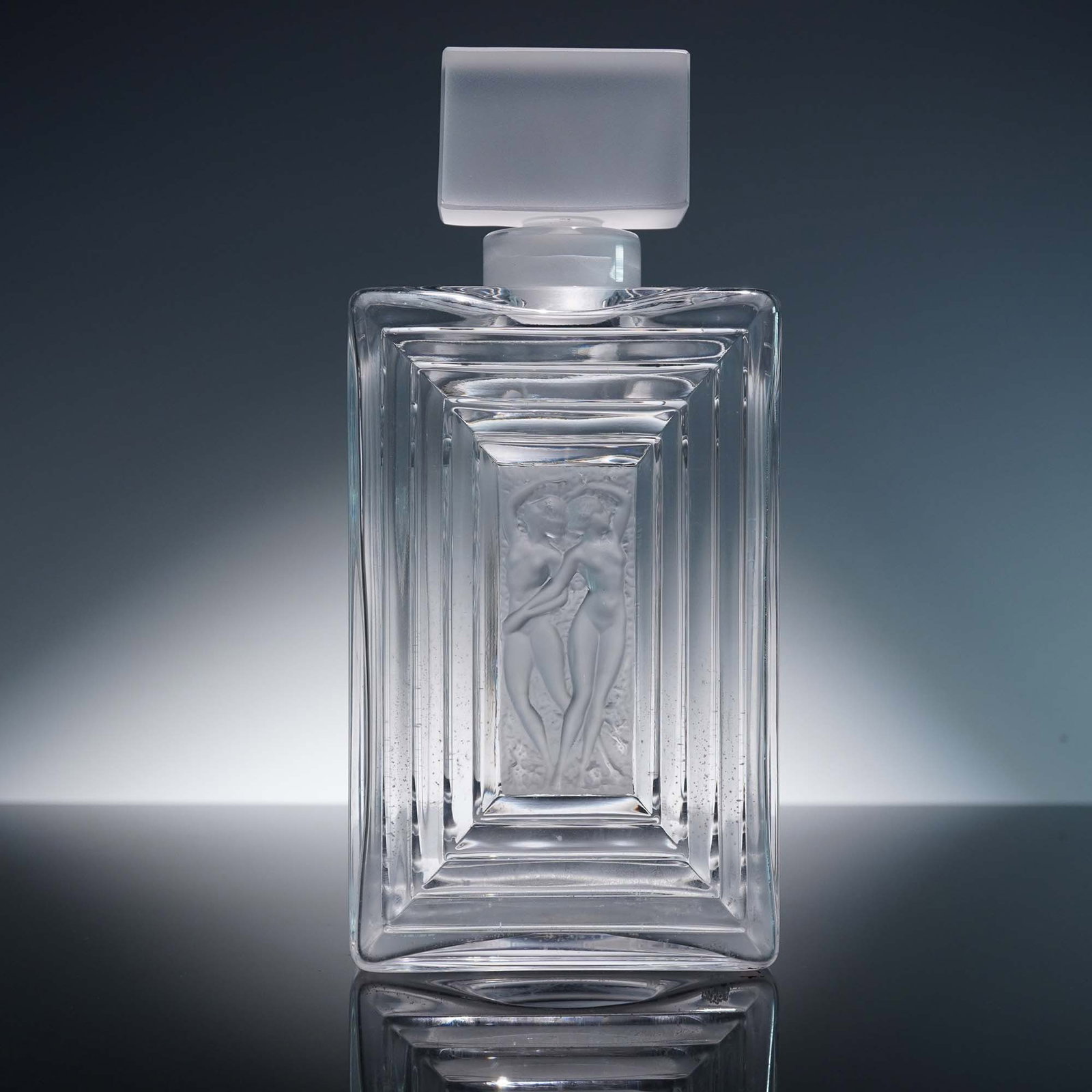 Lalique Crystal Perfume Bottle, Duncan No. 3: A crystal perfume bottle with stopper featuring a bold rectangular silhouette with deeply stepped sides and a central frosted relief of two nude female figures in embrace. This striking Art Deco inspi