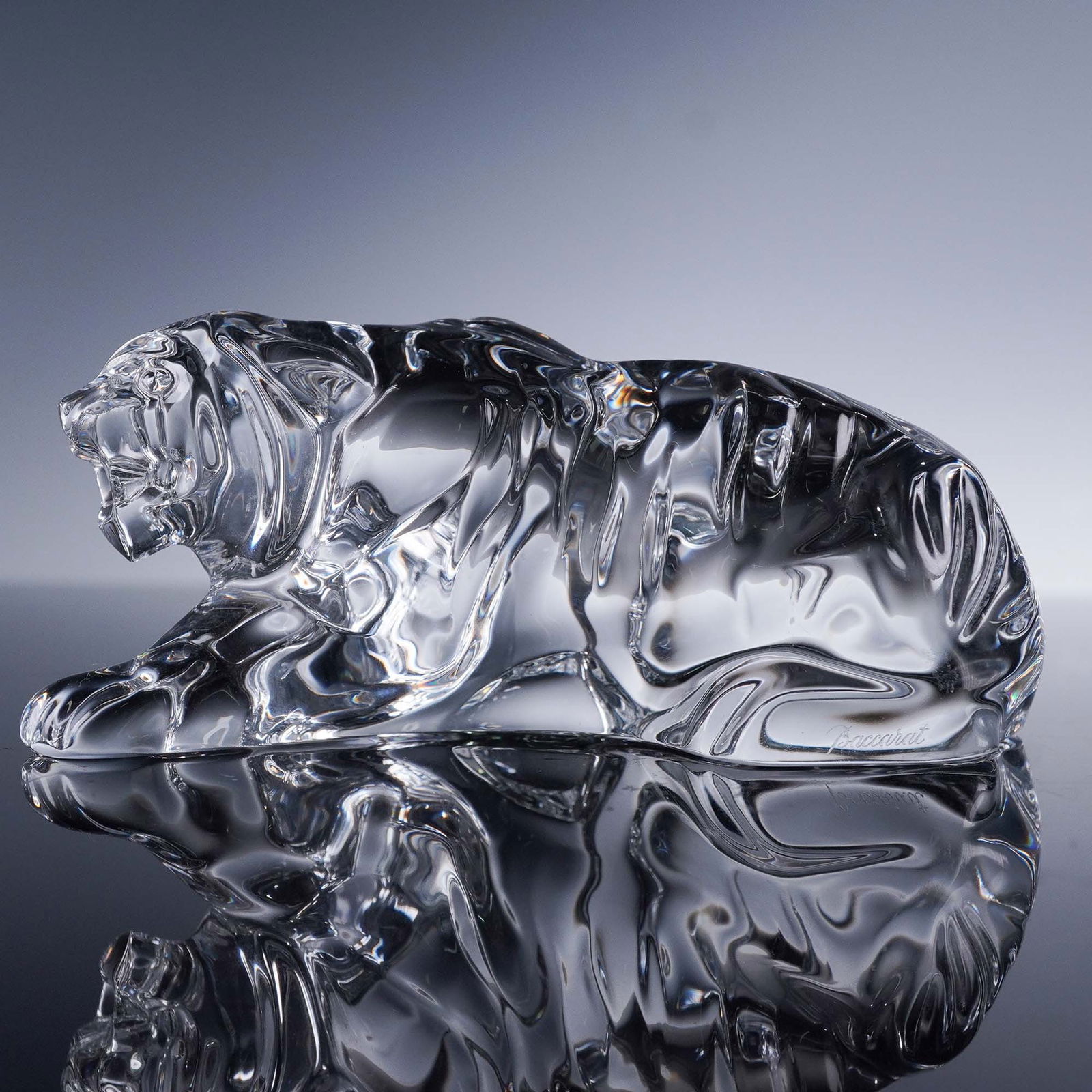 Baccarat Crystal Tiger Figurine: The figure depicts a stylized tiger in a reclining pose with one front paw extended and its mouth open, revealing teeth and a partially turned head. The form incorporates sweeping curves and defined c