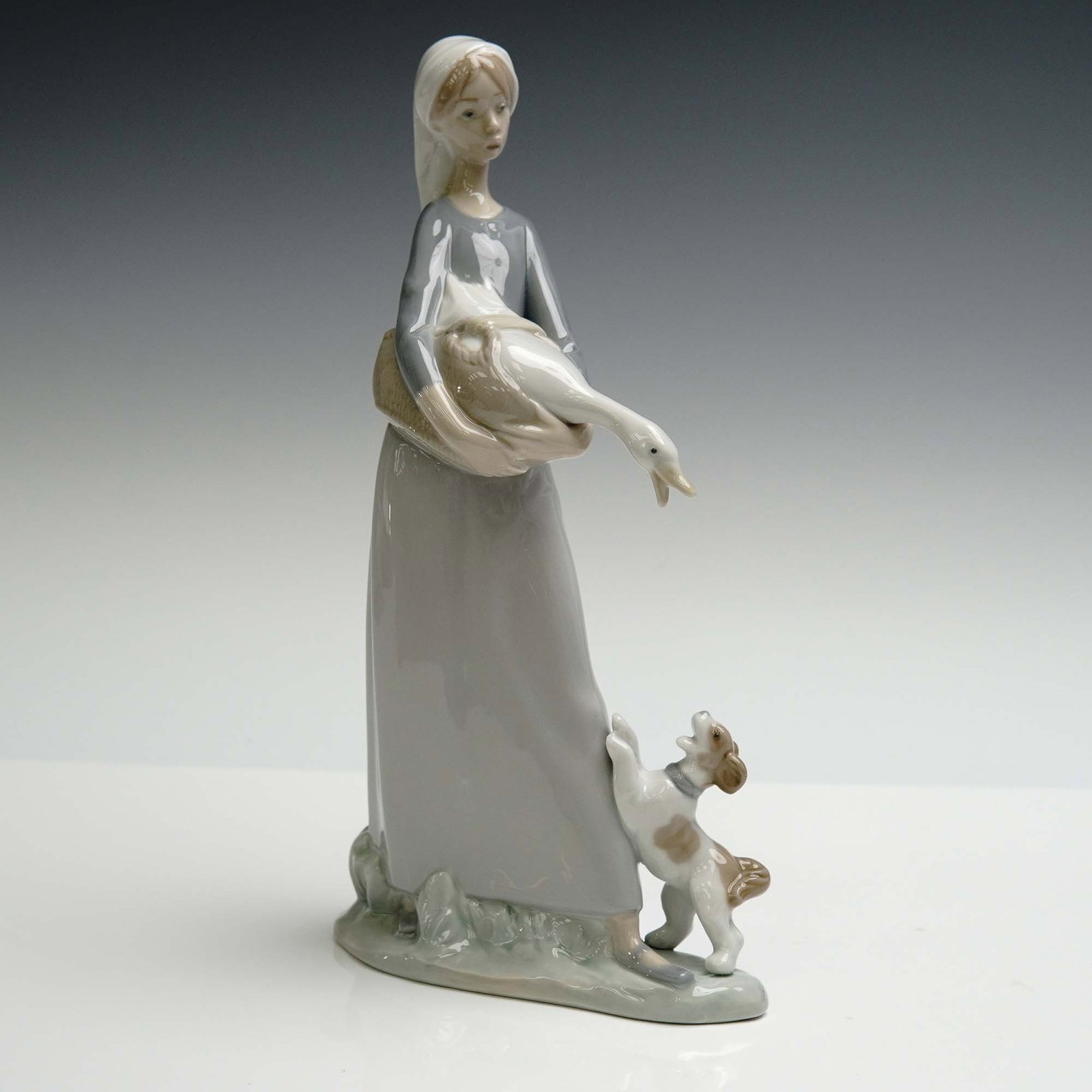 Lladro Porcelain Figurine, Girl W/ Goose and Dog 1008466: Glazed finish featuring a serene young girl gently cradling a goose in her arms while a curious pup reaches up to greet them. Finished in soft gray and earth toned glazes. Lladro backstamp. Artist: Fu