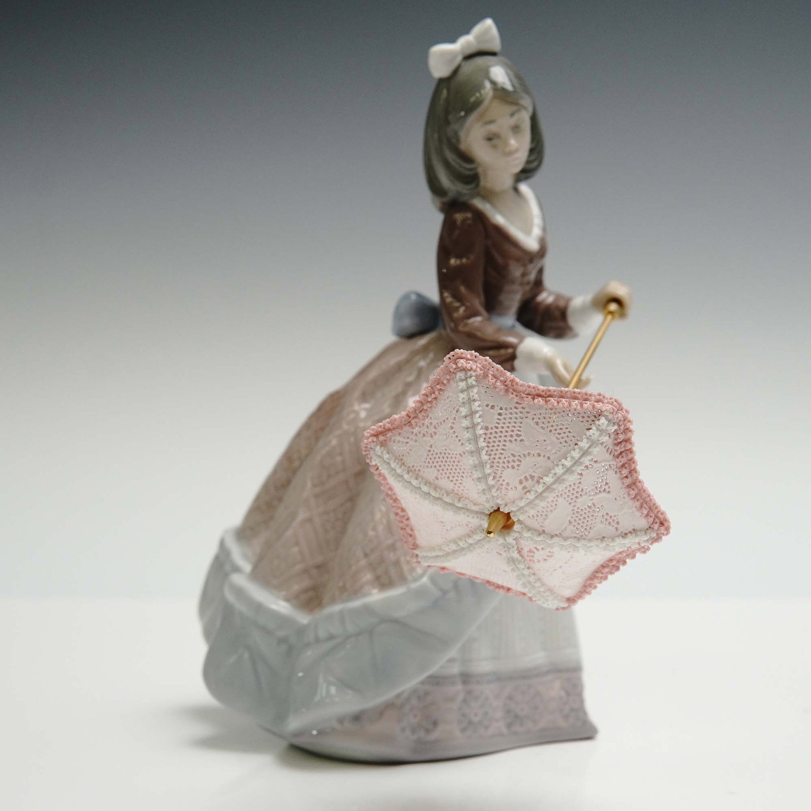 Lladro Porcelain Figurine, Jolie 1005210: Glazed finish figure depicting a young girl in a finely textured Victorian style gown holding a delicately crafted parasol. Lladro backstamp. Artist: Jose Puche Issued: 1984 - 2002 Dimensions: 7"L x 4