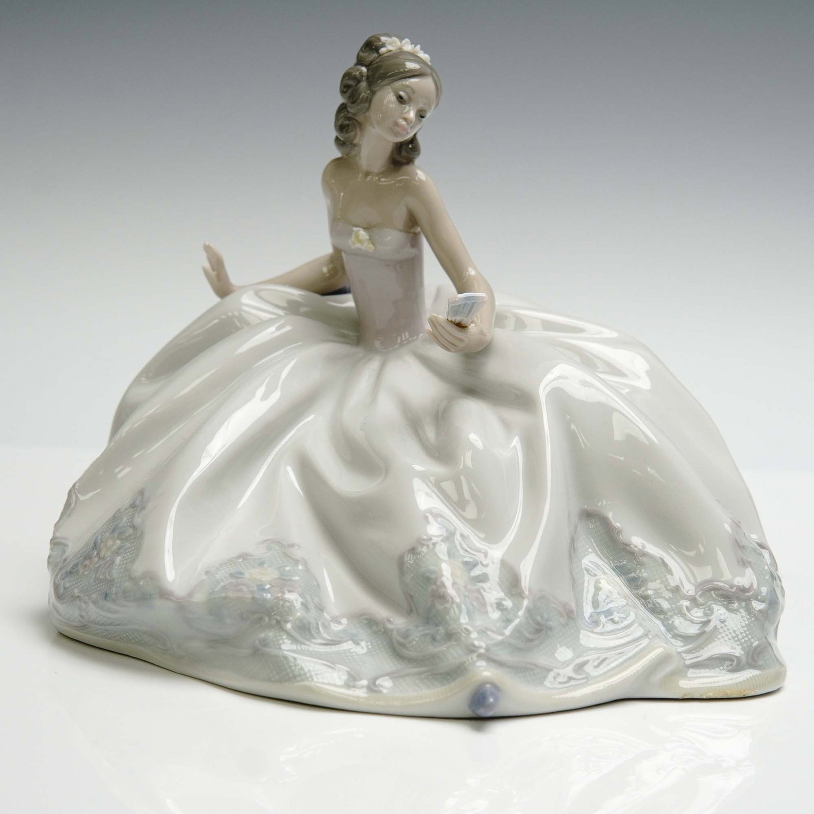 Lladro Porcelain Figurine, At The Ball 1005859: Glazed finish figure depicting a young elegant girl ready for the ball dance wearing a white dress with patten at the ends, holding a folding fan. Lladro backstamp. Artist: Jose Puche Issued: 1992 Dim