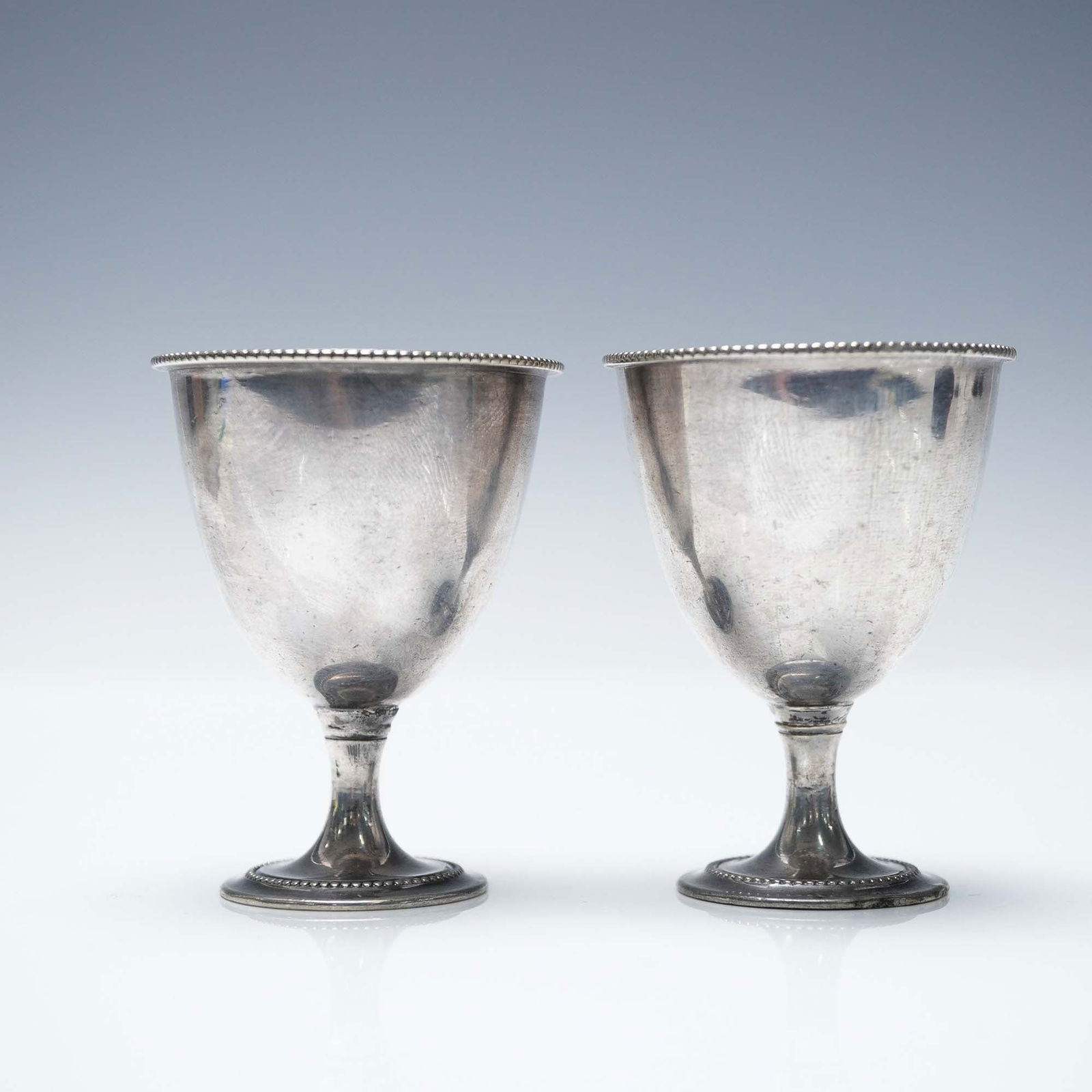 2pc Silver Plated Egg Cups: A pair of silver-plated egg cups featuring a classic beaded rim and pedestal base design. Each cup measures 2.5"H x 2" in diameter. The interiors have a subtle gold wash, adding a touch of elegance. A