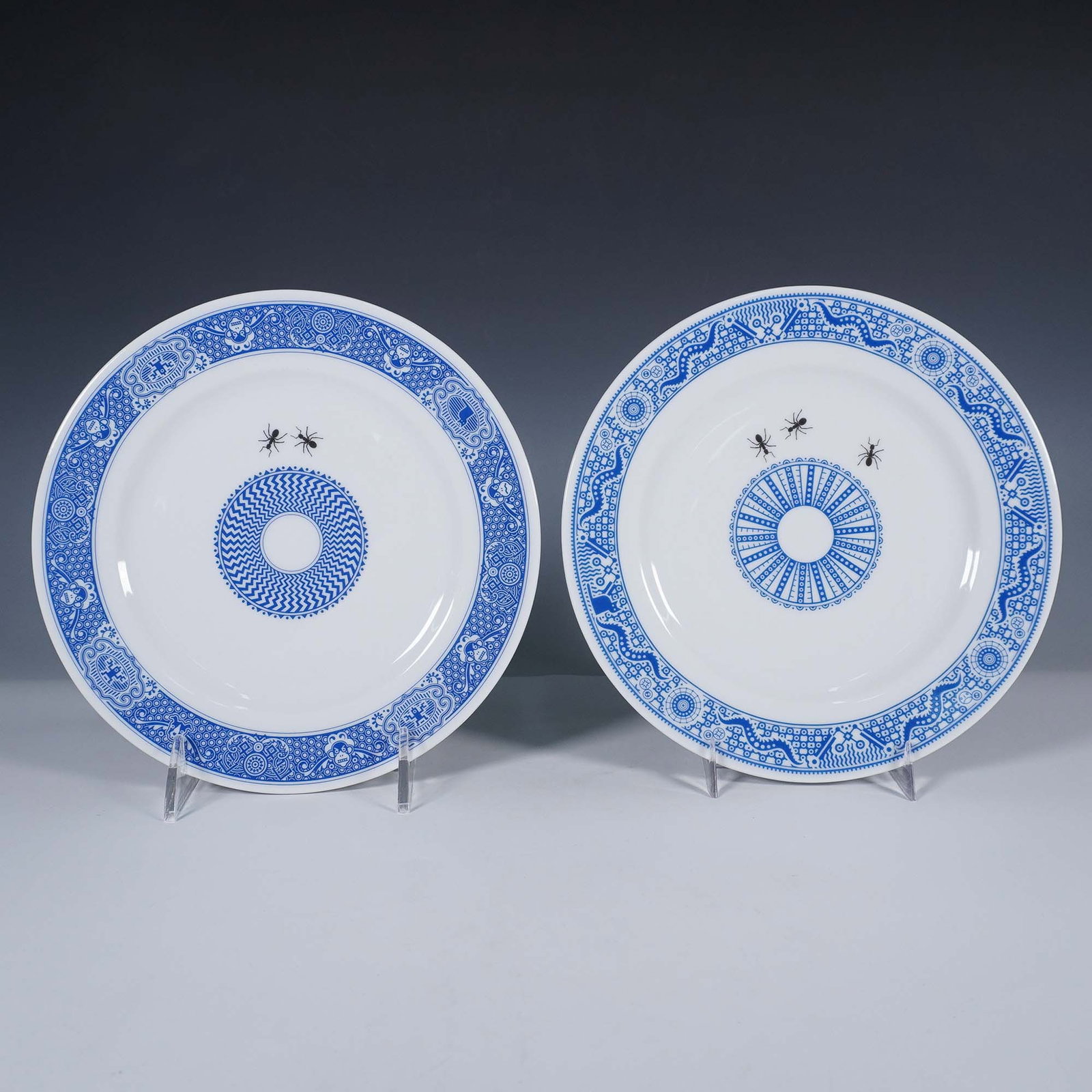 2pc Calamityware Small Plates, Ants: Features 2 small plates with a blue geometric pattern on a white base. The rim includes hidden images within the decorative motif, such as a cat and frowning face. The base includes a circular geometr
