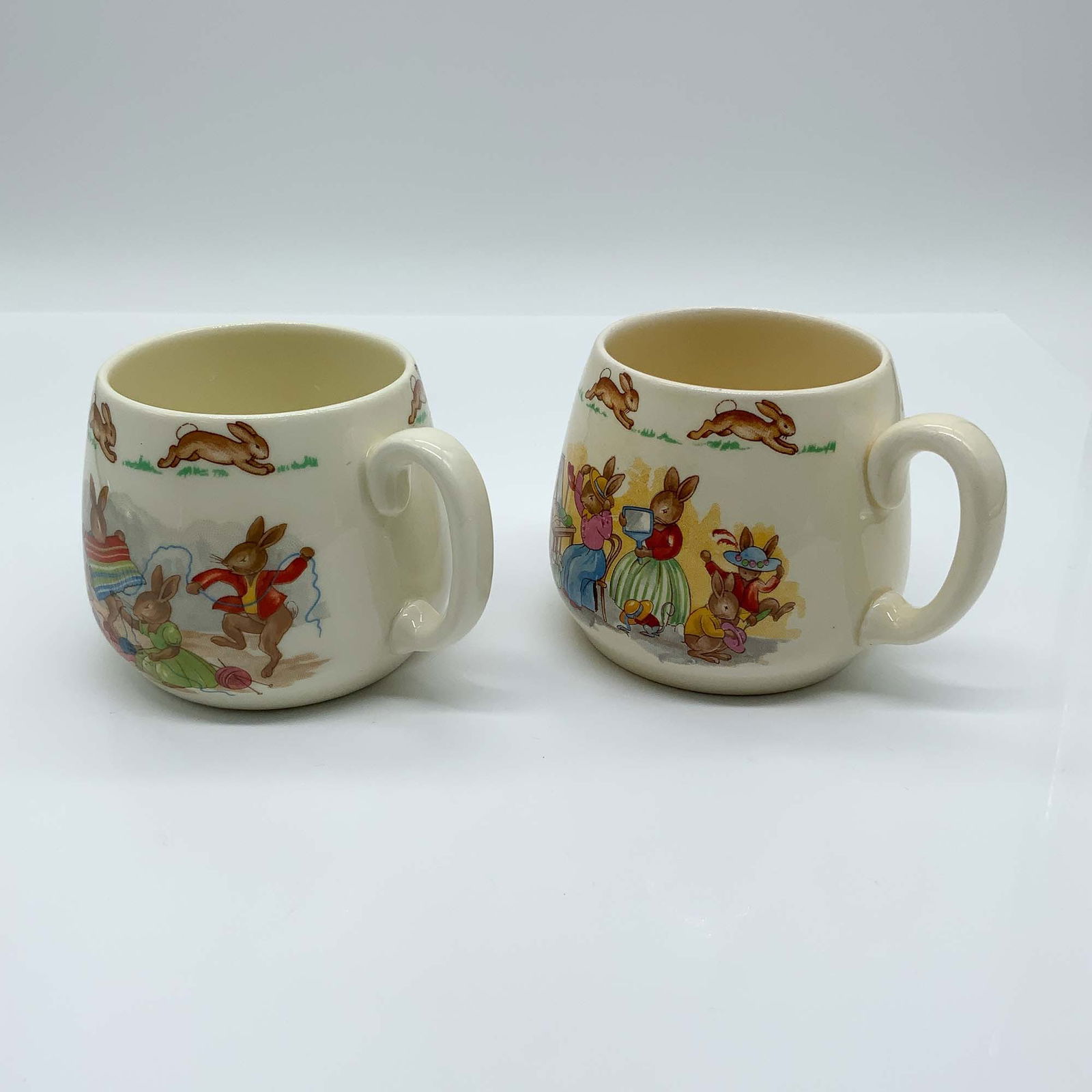 2pc Royal Doulton Bunnykins Mugs, Childrens Cups: his lot features a delightful pair of Royal Doulton Bunnykins mugs, each showcasing charming hand-painted scenes of playful rabbits in a variety of activities. These mugs, part of the Bunnykins series