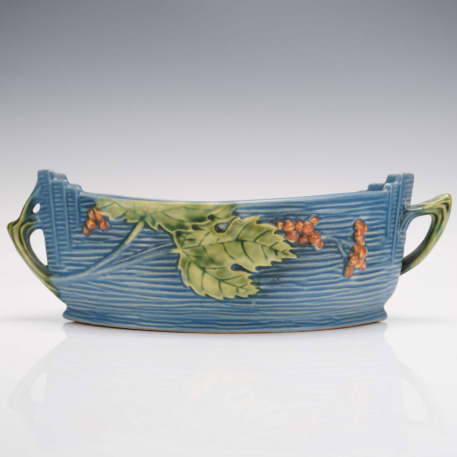 Roseville Pottery Bushberry Blue Console Bowl, 385-10: This Roseville Pottery console bowl in the Bushberry pattern features a rich blue textured background adorned with raised green leaves and clusters of reddish-orange berries. The elongated, canoe-like