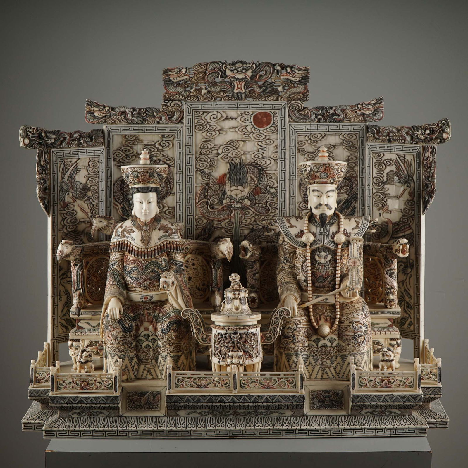 Hand-Carved Bone Polychrome Throne Sculpture, Imperial Emperor & Empress: A refined Chinese hand-carved deer bone sculpture depicting an enthroned imperial couple seated against an intricately carved screen featuring dragons, cranes, clouds, and celestial motifs. The male a