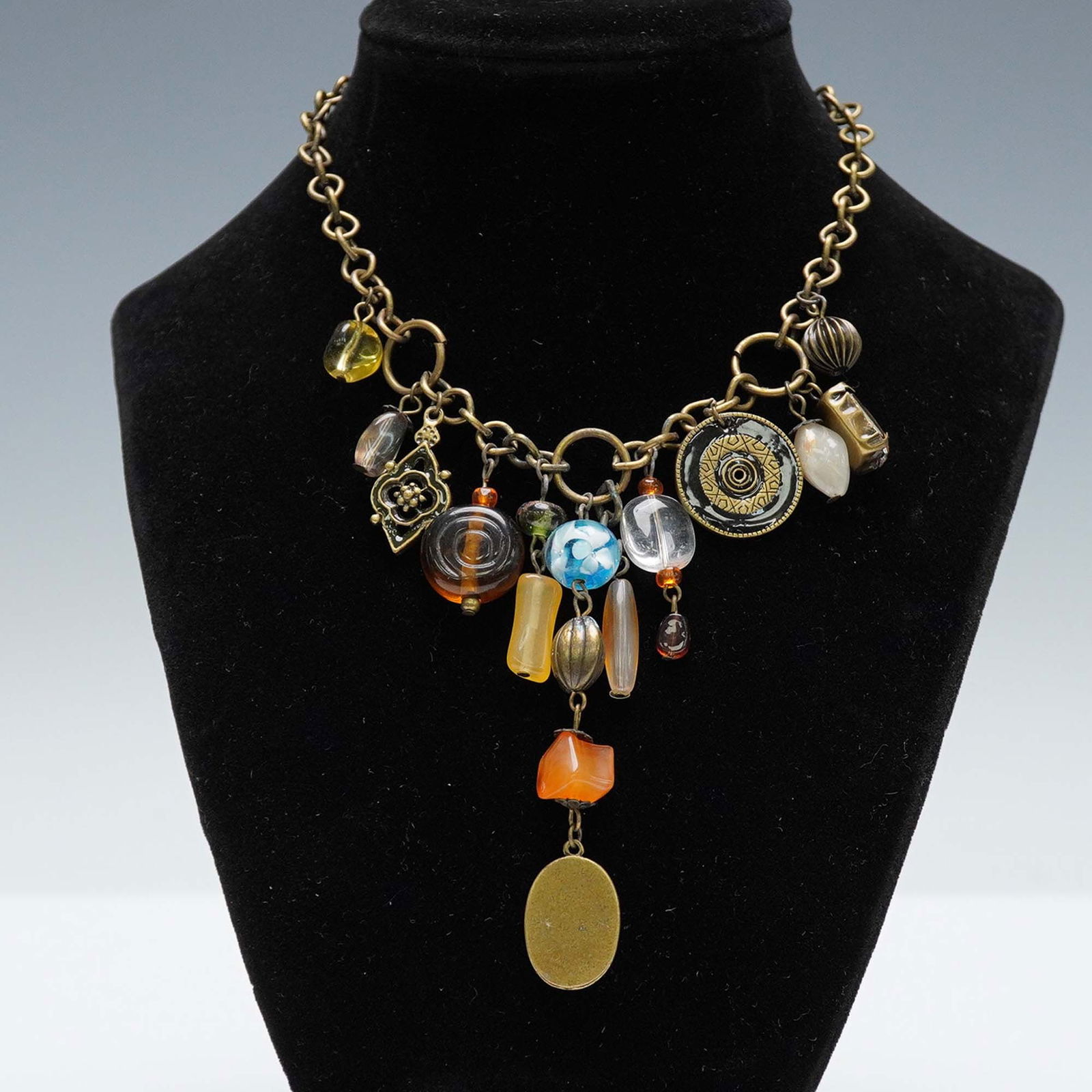 Landau Charm Necklace with Mixed Glass Beads and Dangles: Eclectic charm necklace by Landau featuring a bronze-tone chain adorned with a variety of decorative drops including sculpted glass beads, metal medallions, and enamel accents. The center is finished