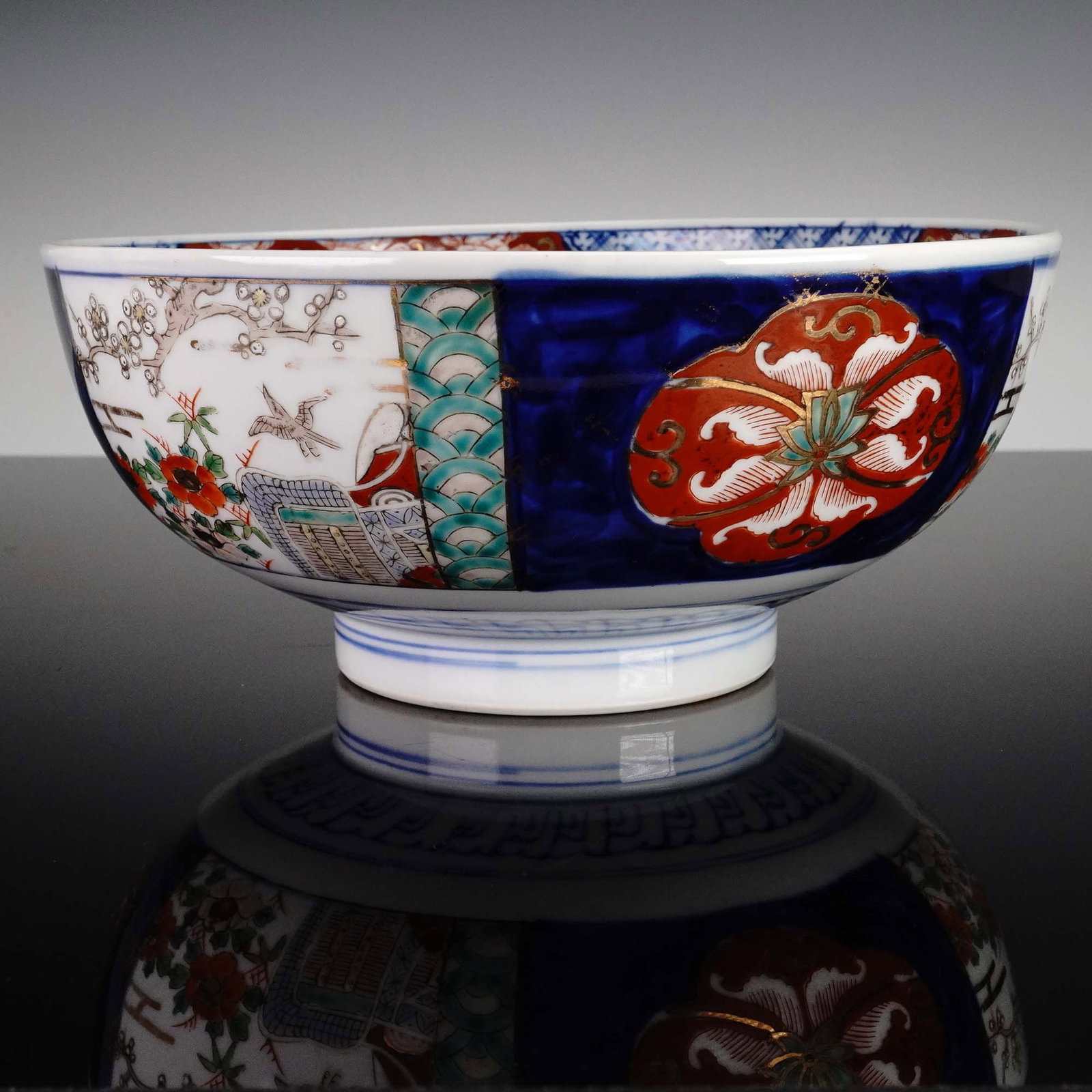 Japanese Porcelain Bowl, Arita Ware in Imari Style: Japan: Arita, mid-20th century. This finely crafted porcelain bowl exemplifies the vibrant Imari style of Arita ware, featuring alternating scenic and floral panels in cobalt blue, red, and emerald gr