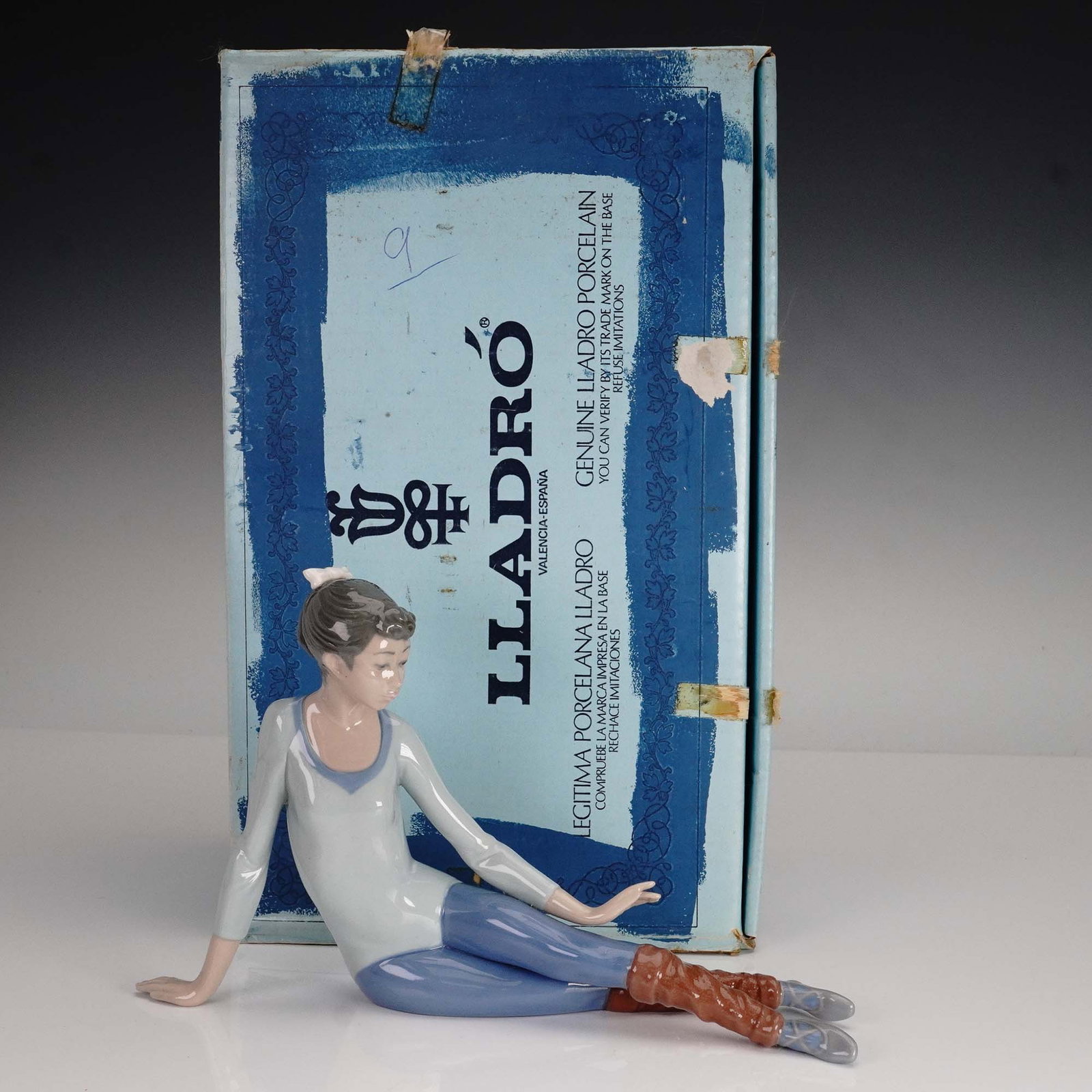 Lladro Porcelain Figurine, Aerobics Scissor 5336: Crafted by the renowned Spanish porcelain house Lladro, this figurine titled Aerobics Scissor captures a young dancer in a relaxed seated pose, dressed in a pale blue leotard and brown leg warmers. Wi